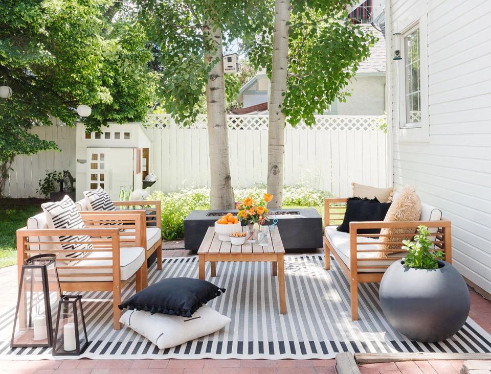 Scandinavian-inspired patio with natural wood seating, striped rug, black accents, and lush greenery overhead