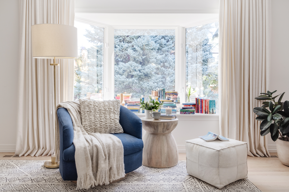 Serene reading nook with blue curved armchair, cream throws, pedestal table, and floor-to-ceiling windows overlooking snowy landscape