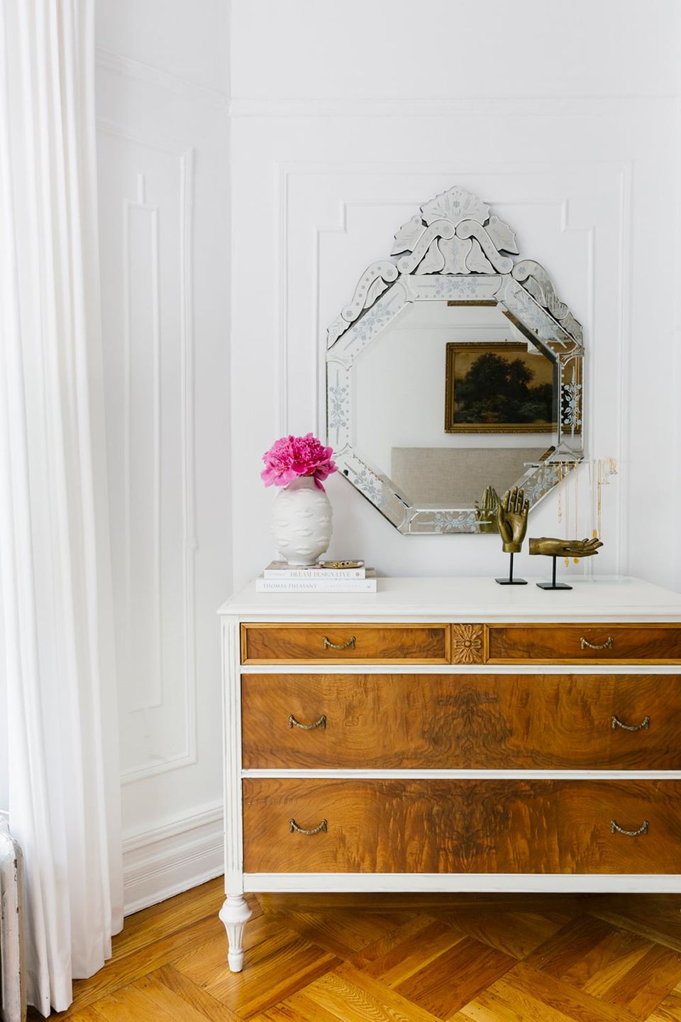 Vintage-modern entryway with ornate mirror, wood dresser, pink florals, and white curtains blending eclectic charm with elegance.