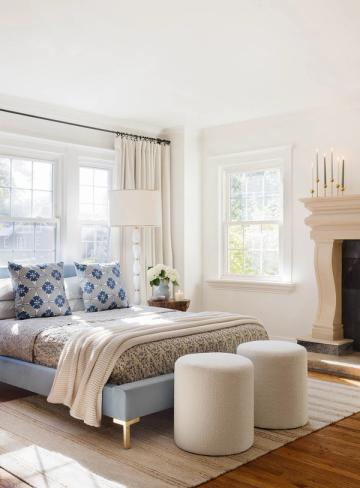 Bright transitional bedroom with blue patterned pillows, cream linens, fireplace, and natural light emphasizing timeless coastal style.