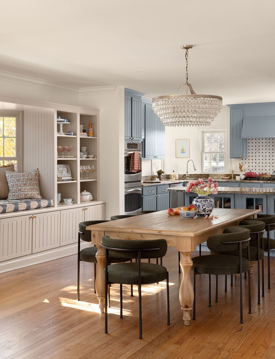 Transitional kitchen-dining blend with soft blue cabinetry, white shelving, natural wood table, and crystal chandelier