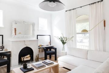Bright Parisian living room with cream sofa, black accents, fireplace, and flowing white curtains creating an elegant, minimalist French aesthetic