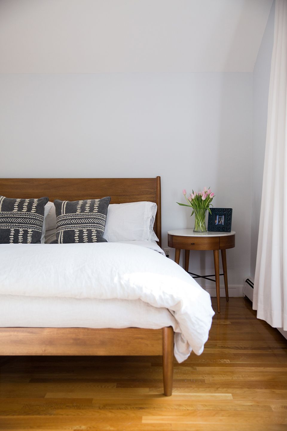 Minimalist mid-century bedroom with walnut wood bed, geometric black pillows, white walls, and natural light.
