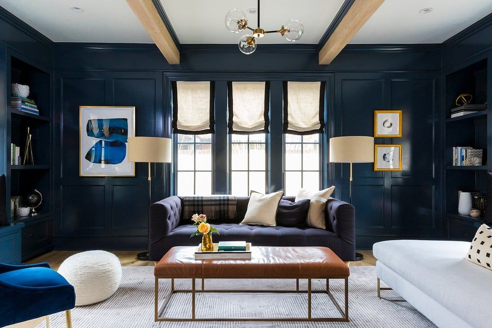 Modern living room with deep navy walls, exposed wood beams, and bright windows creating a sophisticated, moody aesthetic with warm wood furniture.