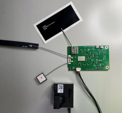 Top-down view of an electronic air quality monitoring prototype on a grey surface. A green printed circuit board is connected by cables to a rectangular solar panel, a slim antenna, a small square GPS module, and a black battery pack. The components are laid out separately but wired together, showing the internal hardware of the device