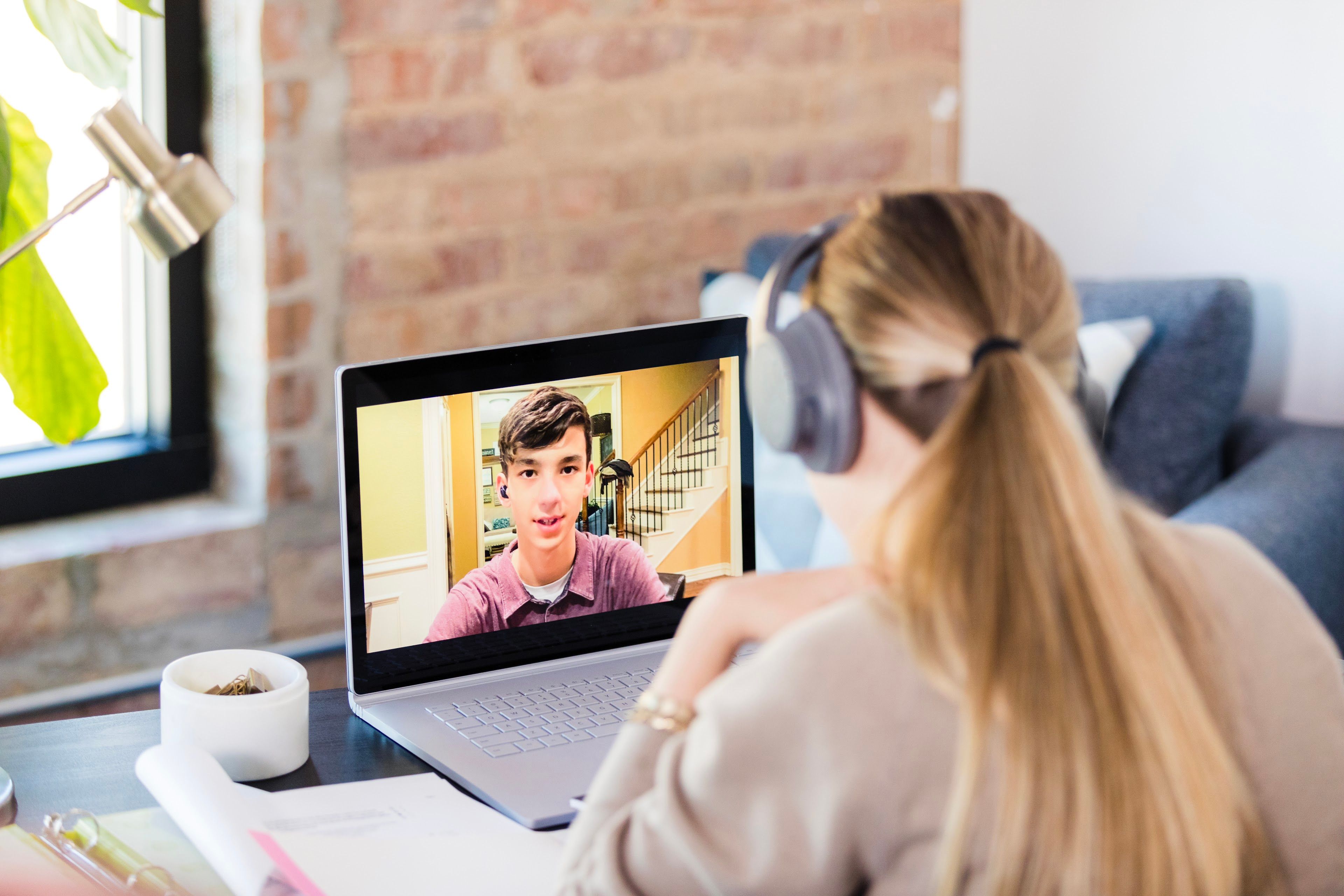 A person wearing headphones watches a young man on a video call on a laptop.