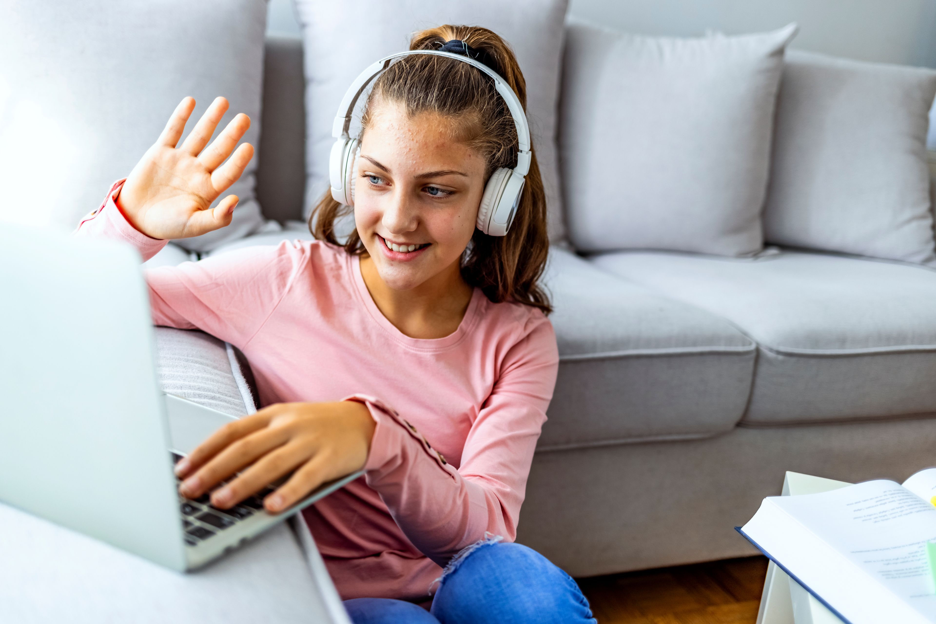 A smiling young girl wearing headphones waves at a laptop screen while sitting on the floor.