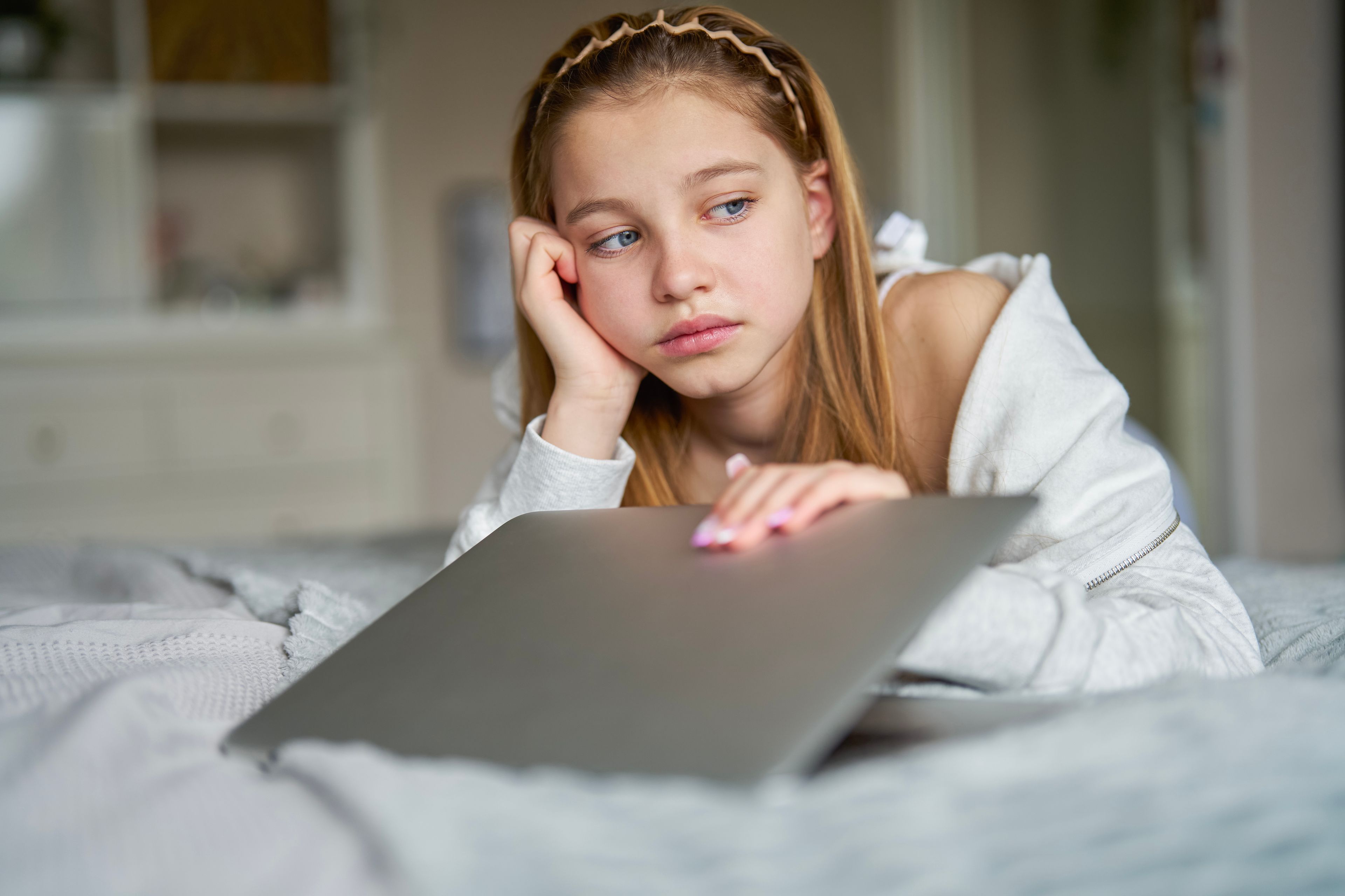 A girl lies on her bed with her head resting on her hand and her laptop closed in front of her.