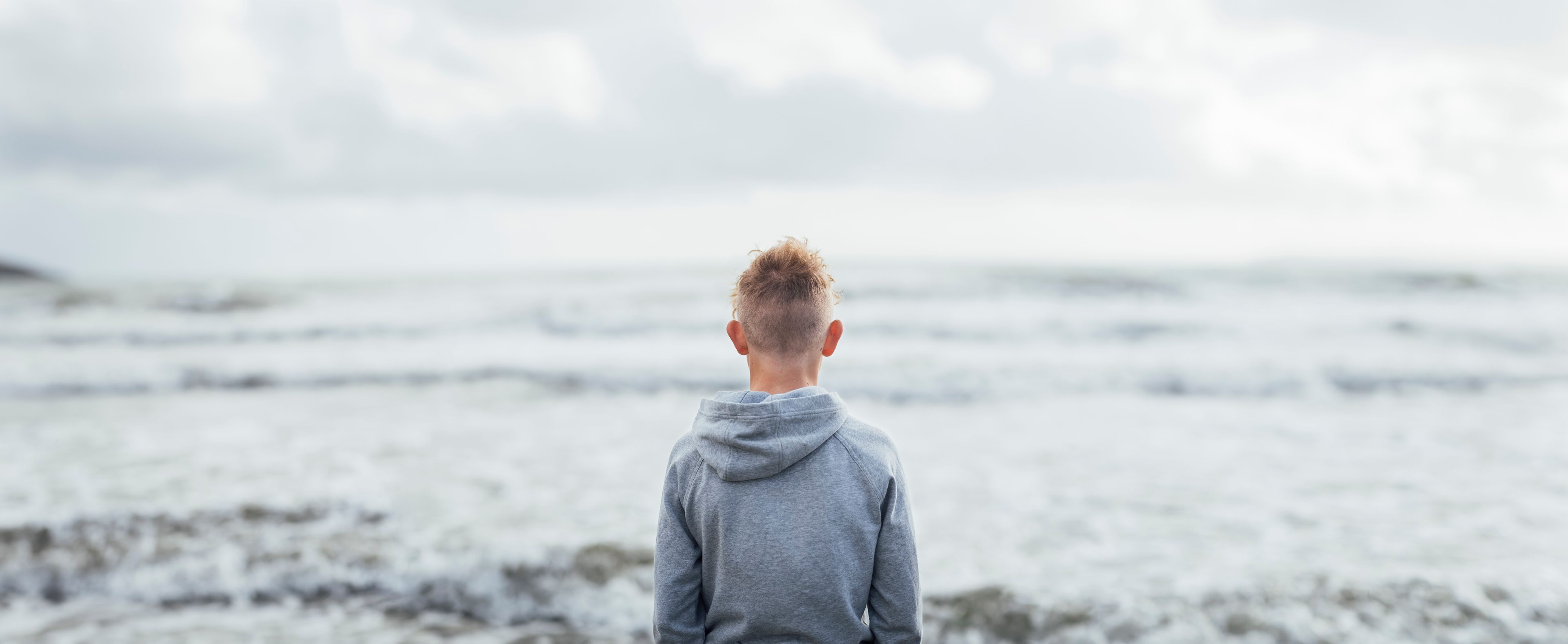 A boy in a grey hoodie, seen from behind, looks out at the ocean.