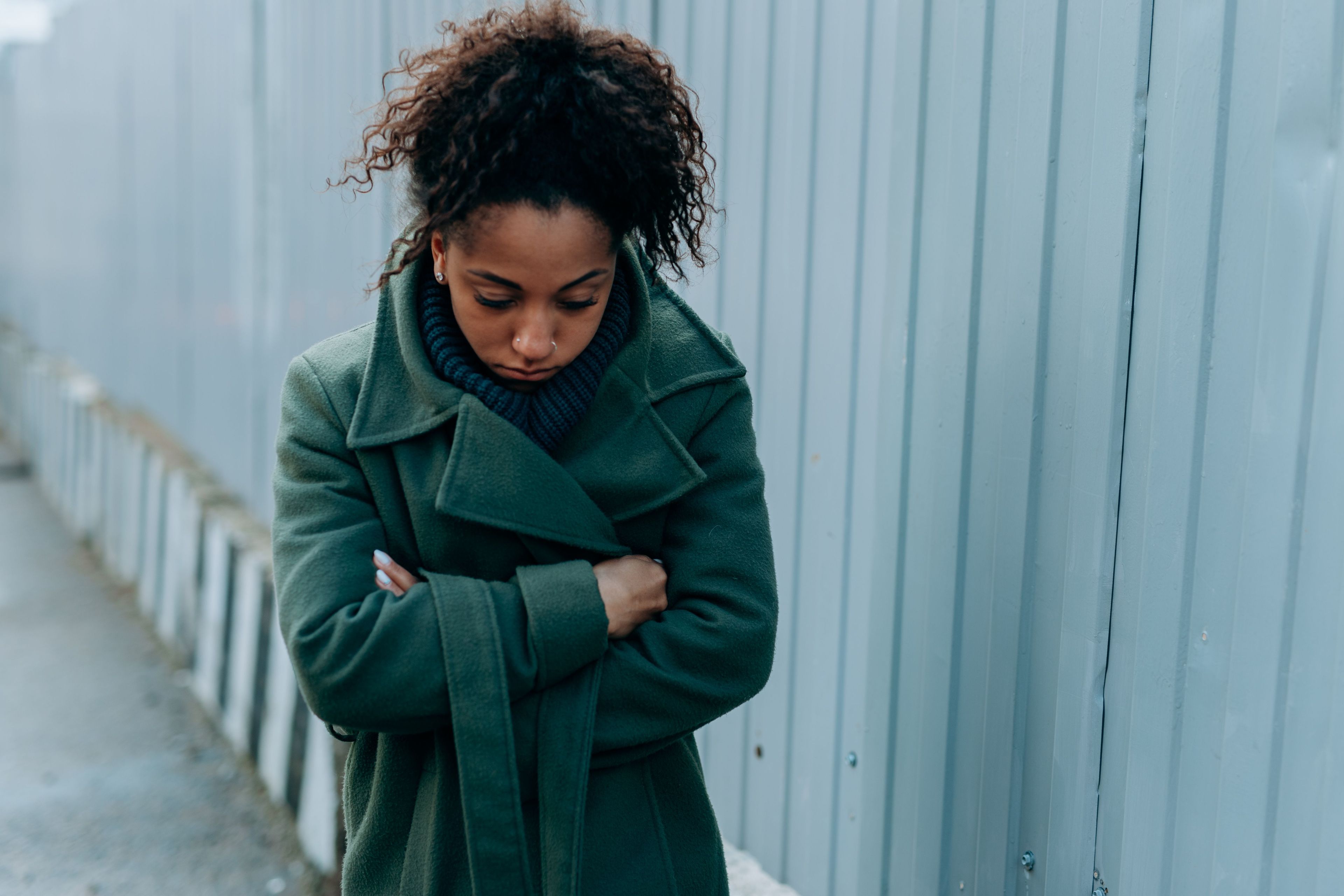 A young woman in a green winter coat stands with her arms folded, looking down, beside a grey metal fence.