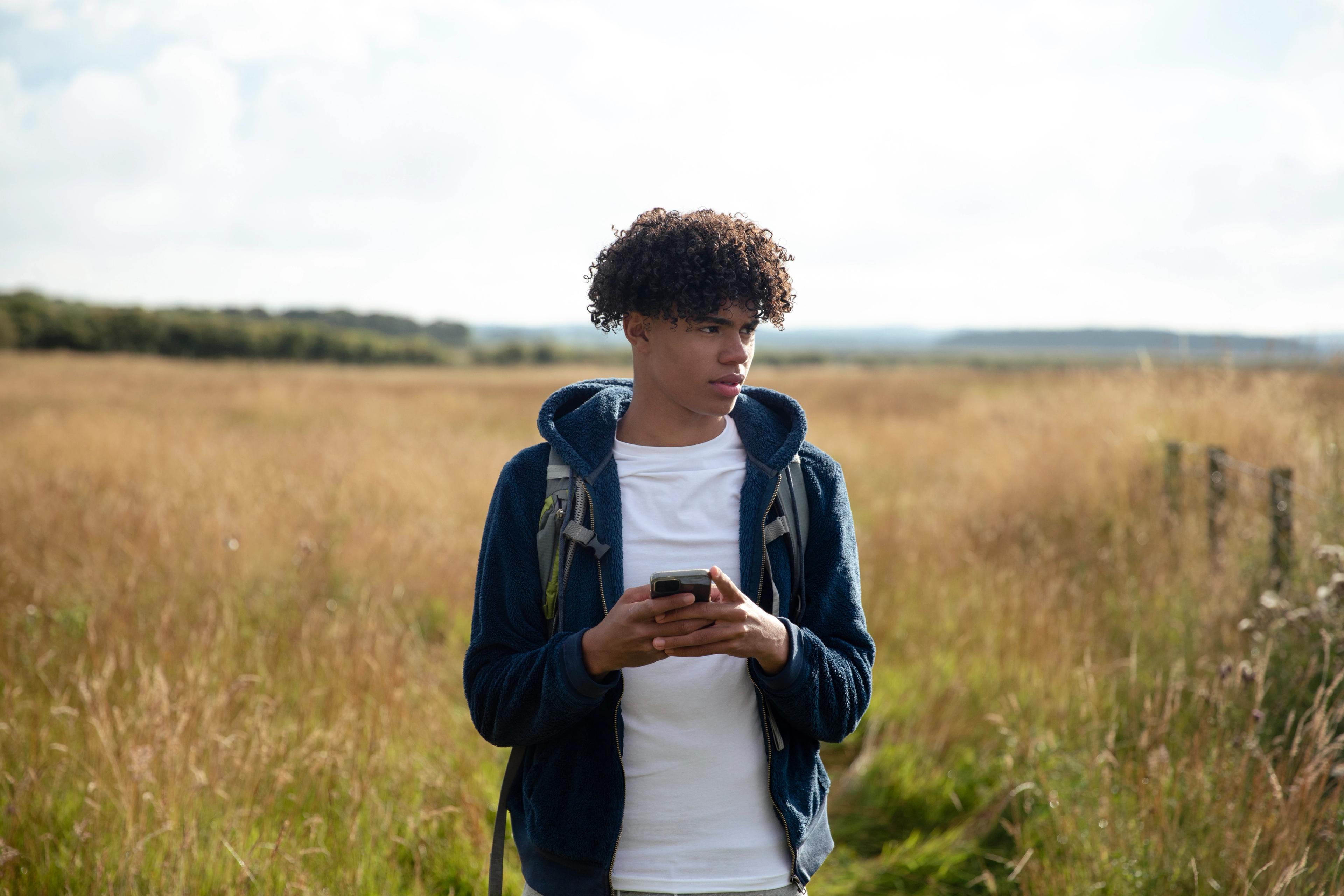 A young man with a backpack holds a phone while walking through a field of tall, dry grass.