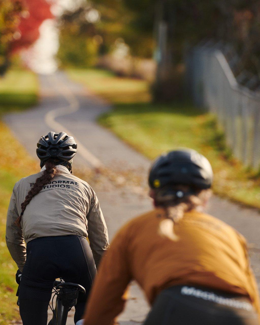 Model wearing beige long-sleeve jersey with back branding, cycling on autumn road with another rider