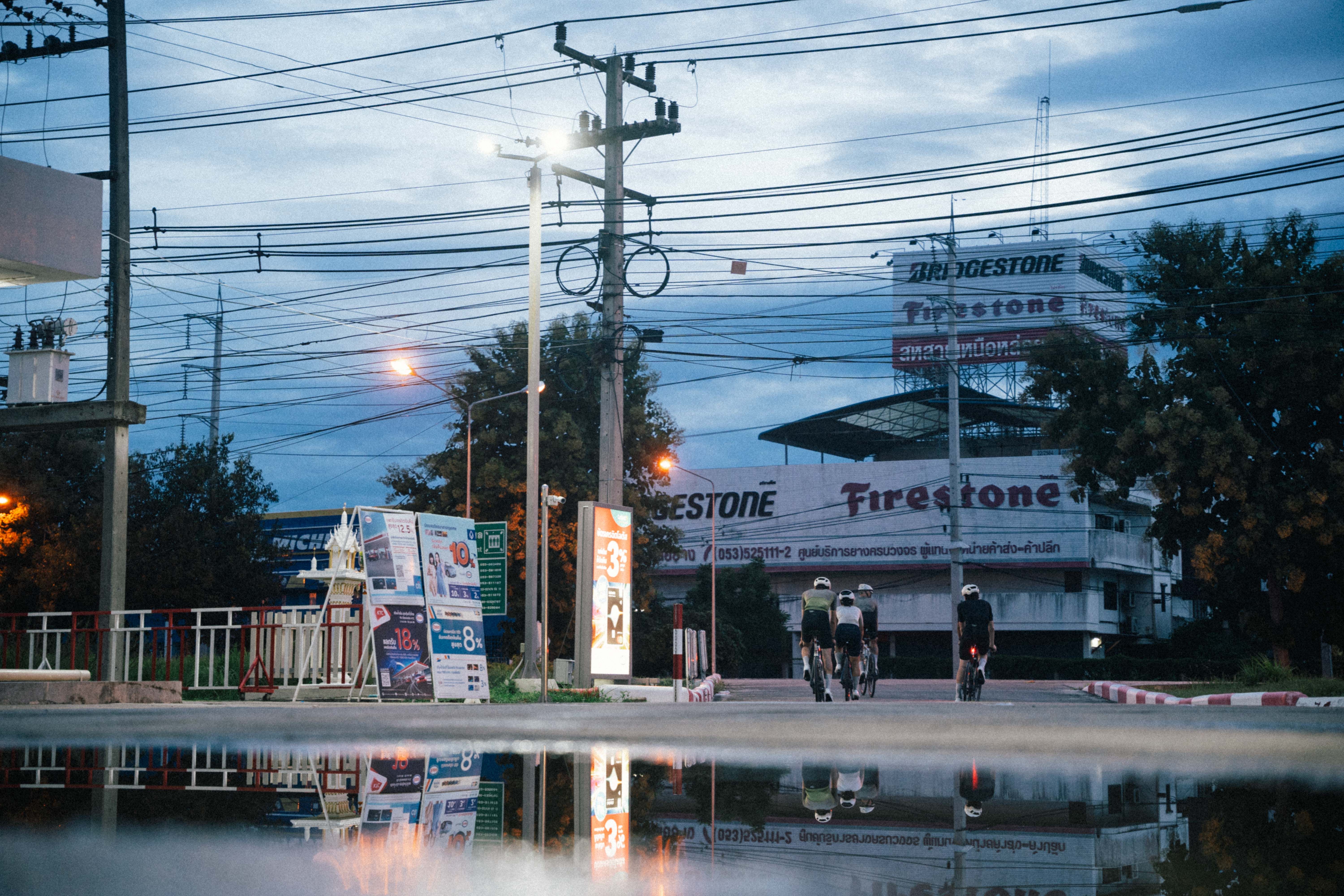 Four cyclists riding through urban Bangkok street at dusk with power lines and Firestone tire shop