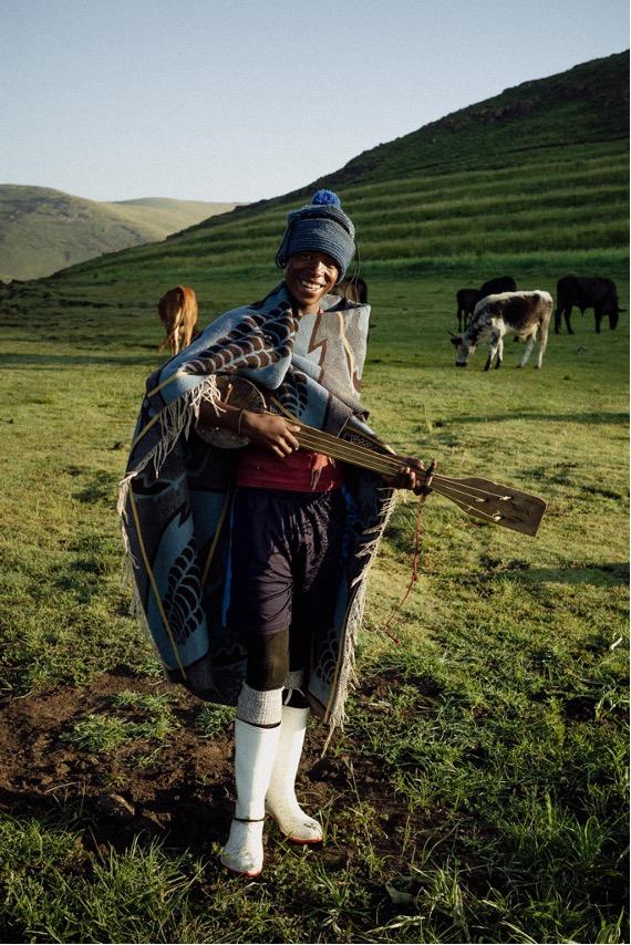Model in blue beanie and patterned blanket holding traditional instrument in mountainous grassland with grazing cattle