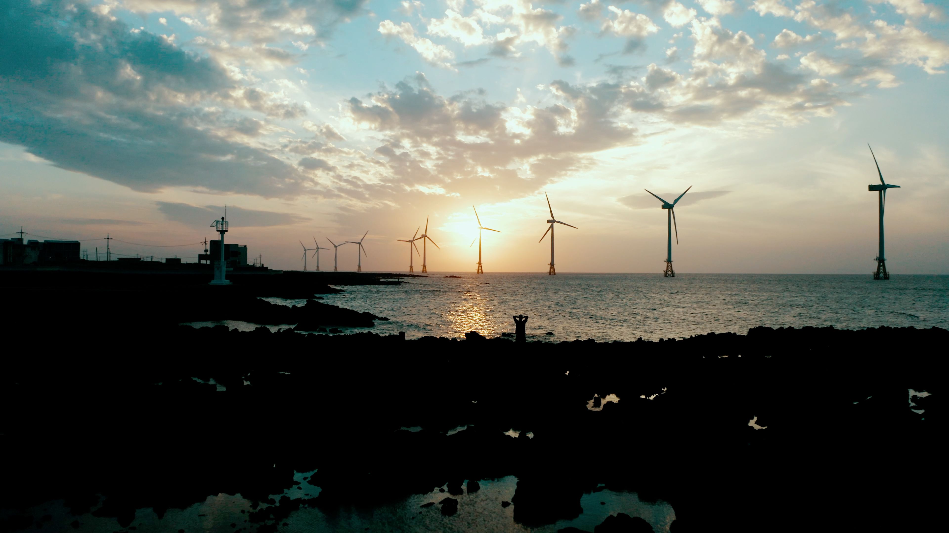 Coastal sunset with offshore wind turbines and silhouetted figure on rocky shoreline