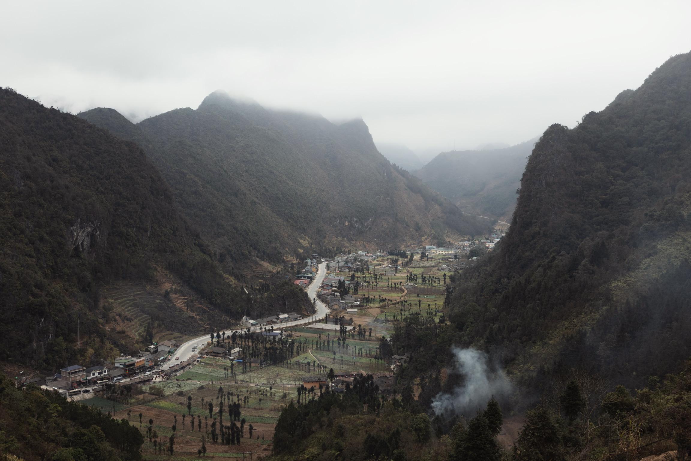 Mountain valley landscape with winding road through small village surrounded by forested hills and misty peaks