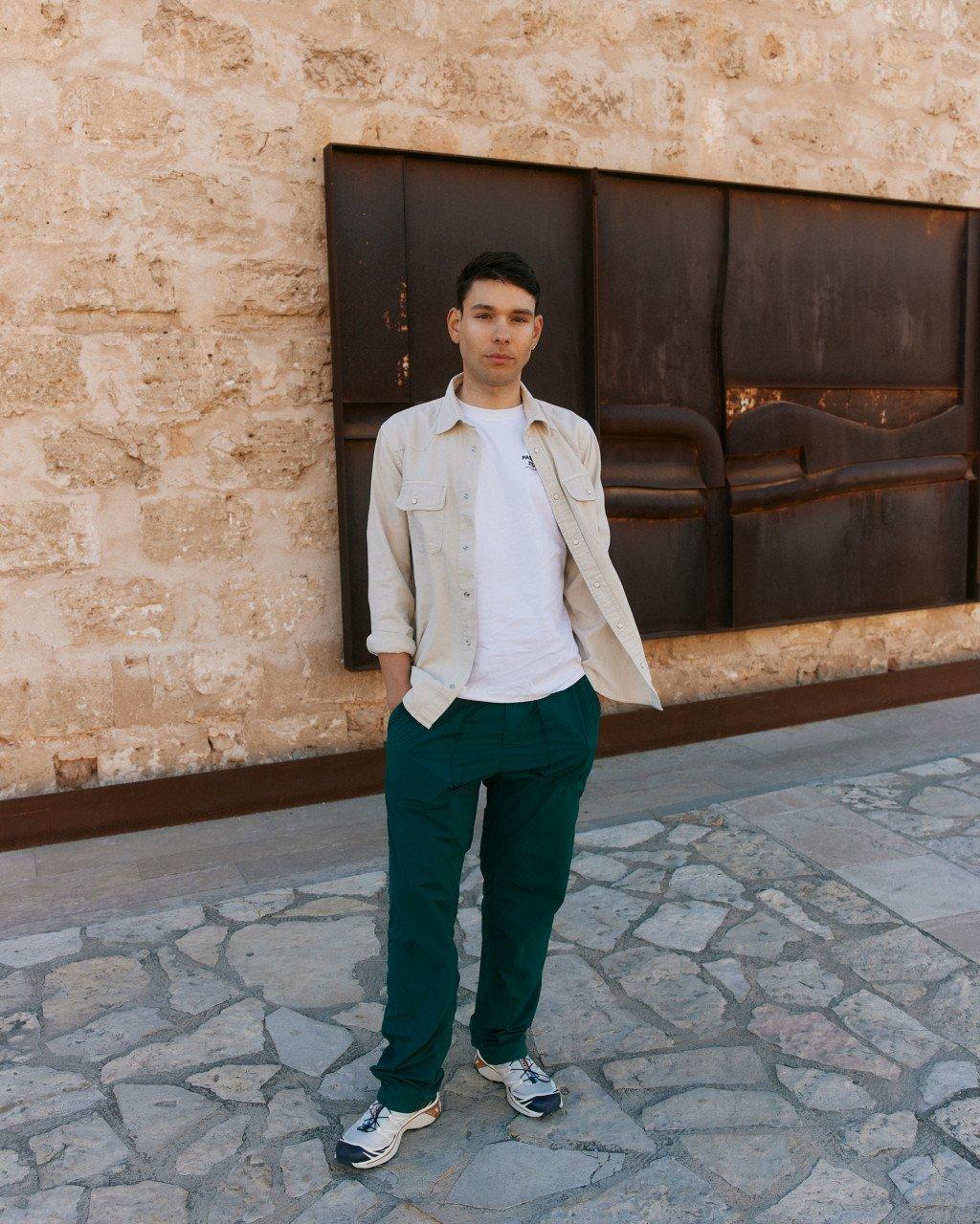 Model wearing white t-shirt and beige shirt with green pants on cobblestone street