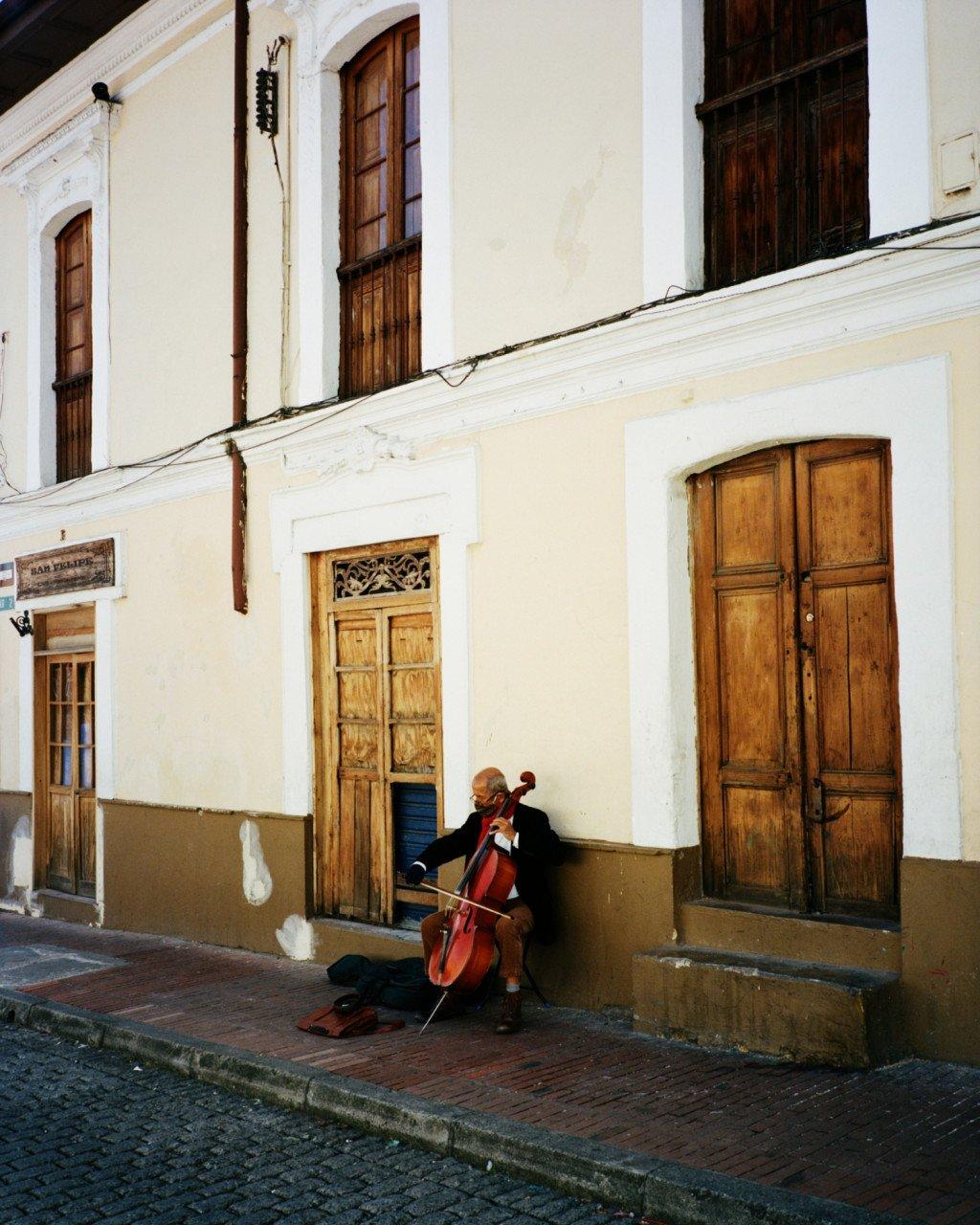 Street musician playing cello outside colonial building with wooden doors and white walls