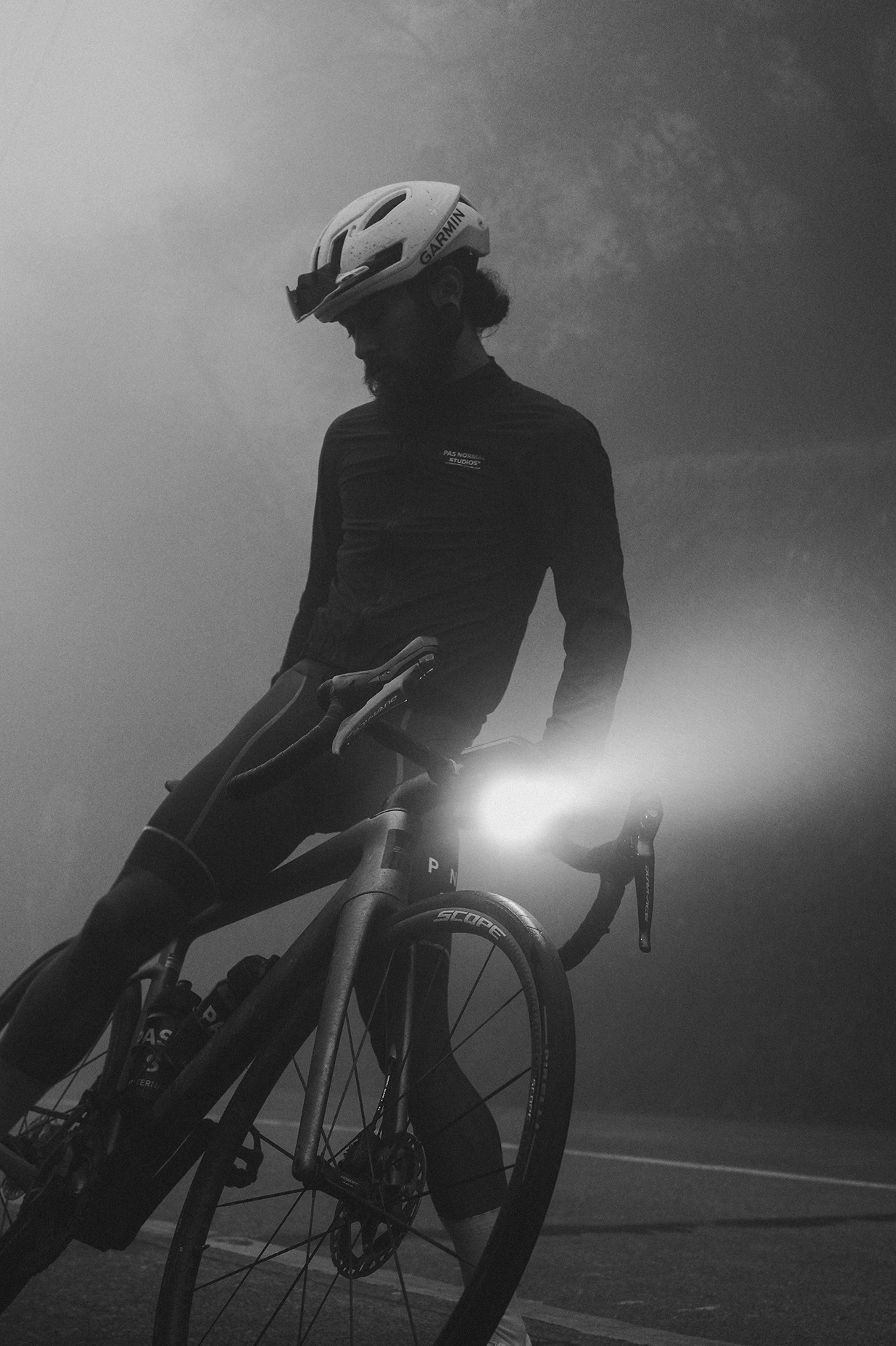 Model wearing dark cycling jersey with helmet, positioned on bike with bright front light in misty conditions