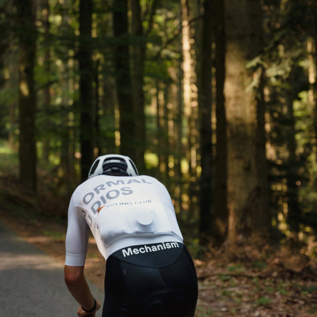Model wearing white Mechanism jersey cycling through wooded area in aerodynamic position