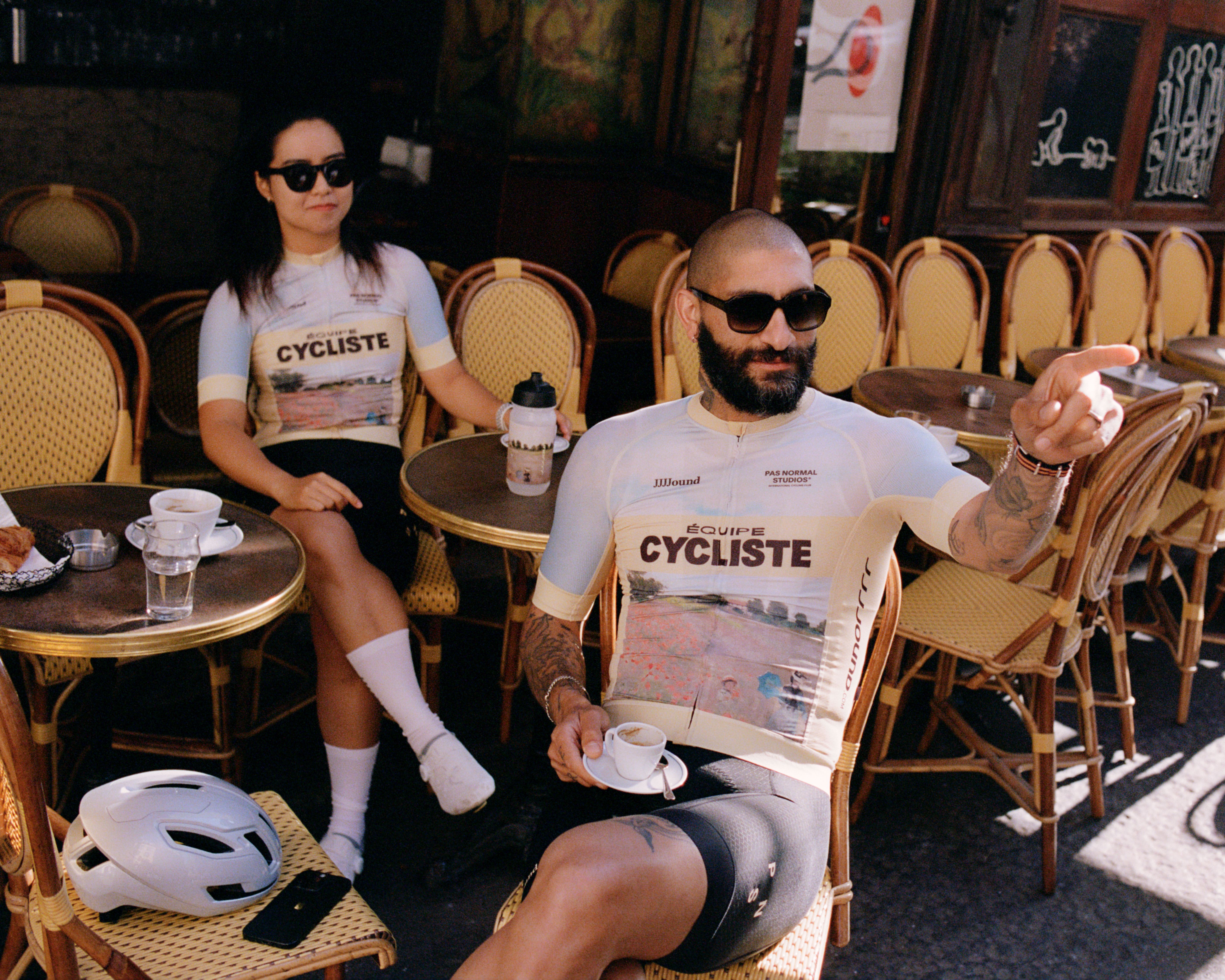 Models wearing white cycling jerseys with Cycliste graphic sitting at Paris café with helmets and coffee