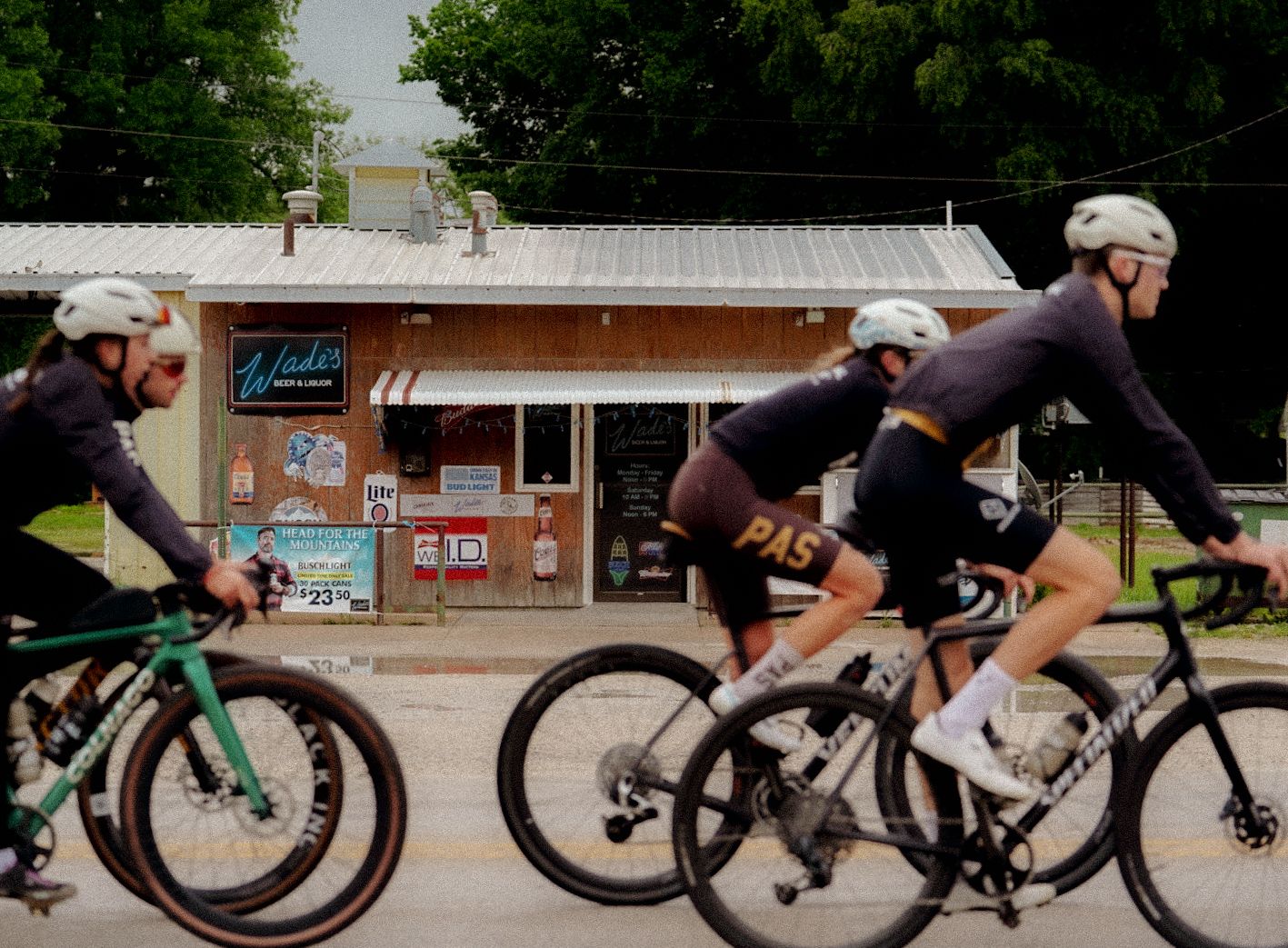 Cyclists racing past rural store wearing black cycling apparel with orange branding details
