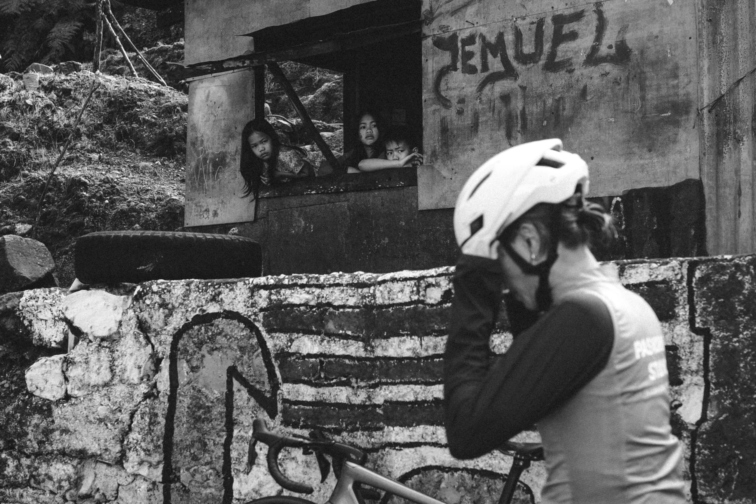 Cyclist in white helmet and cycling apparel on urban street with children watching from building window
