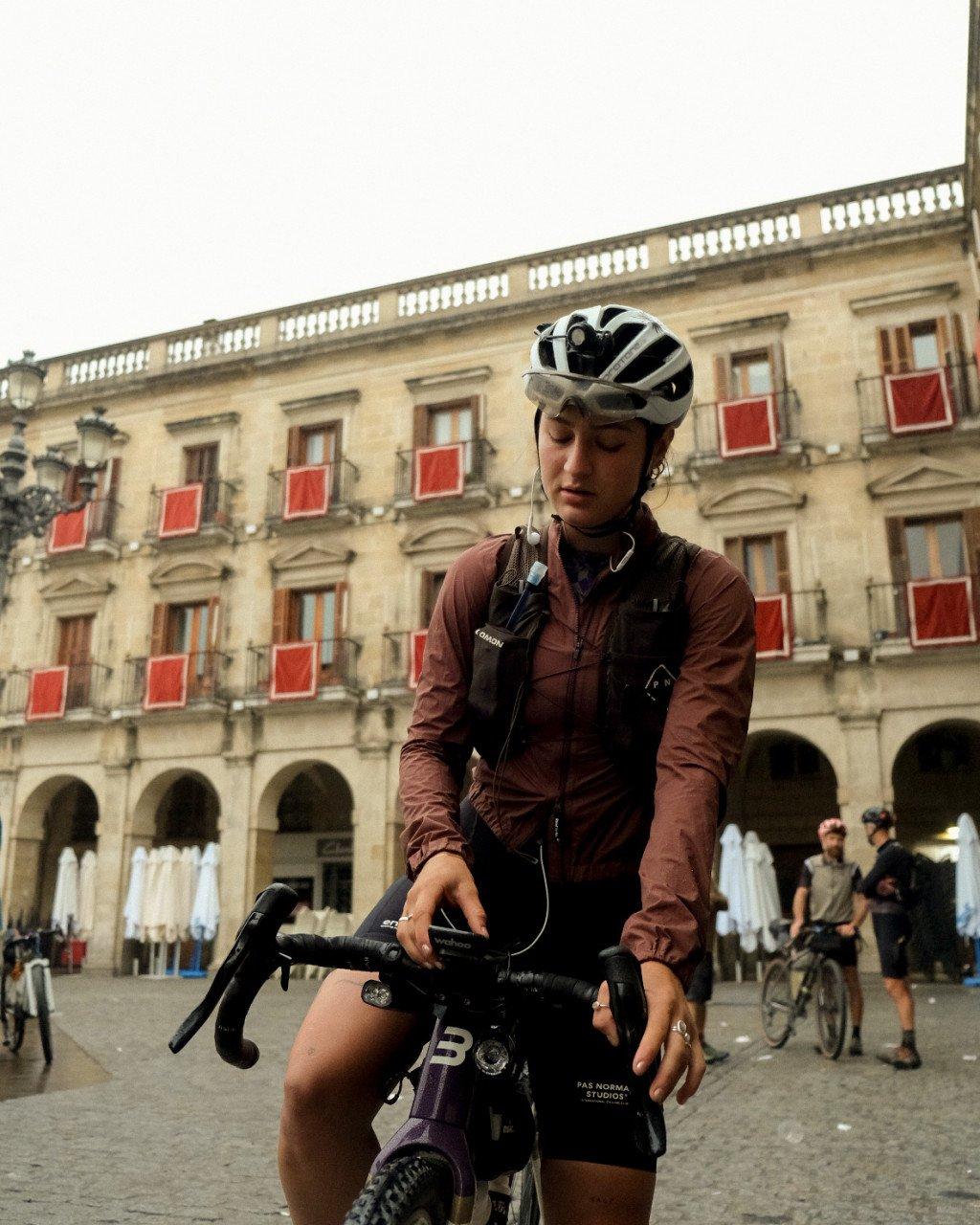 Model wearing brown cycling jacket with helmet and bike in historic town square