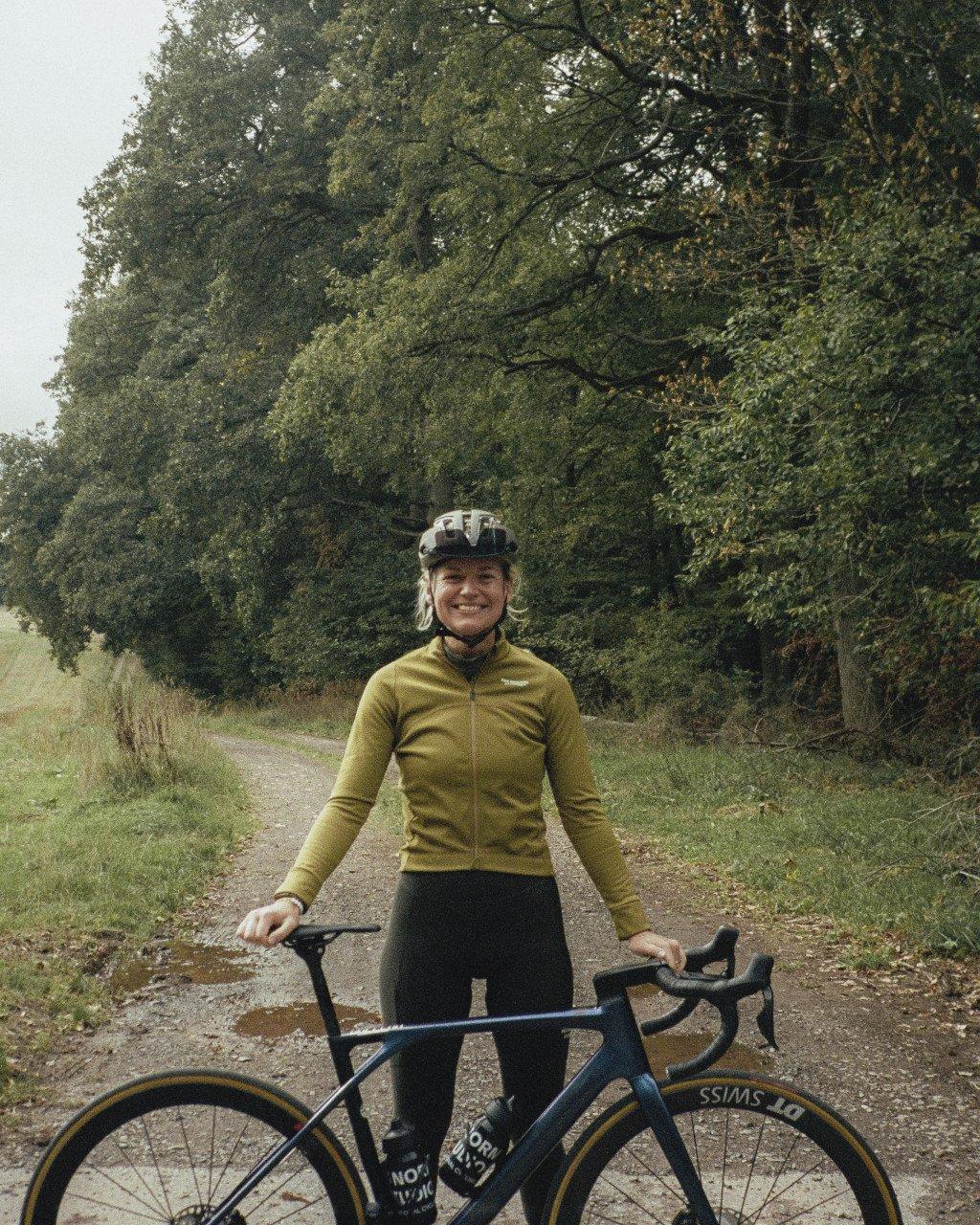 Model wearing yellow long sleeve cycling jersey with white helmet standing with bike on countryside path