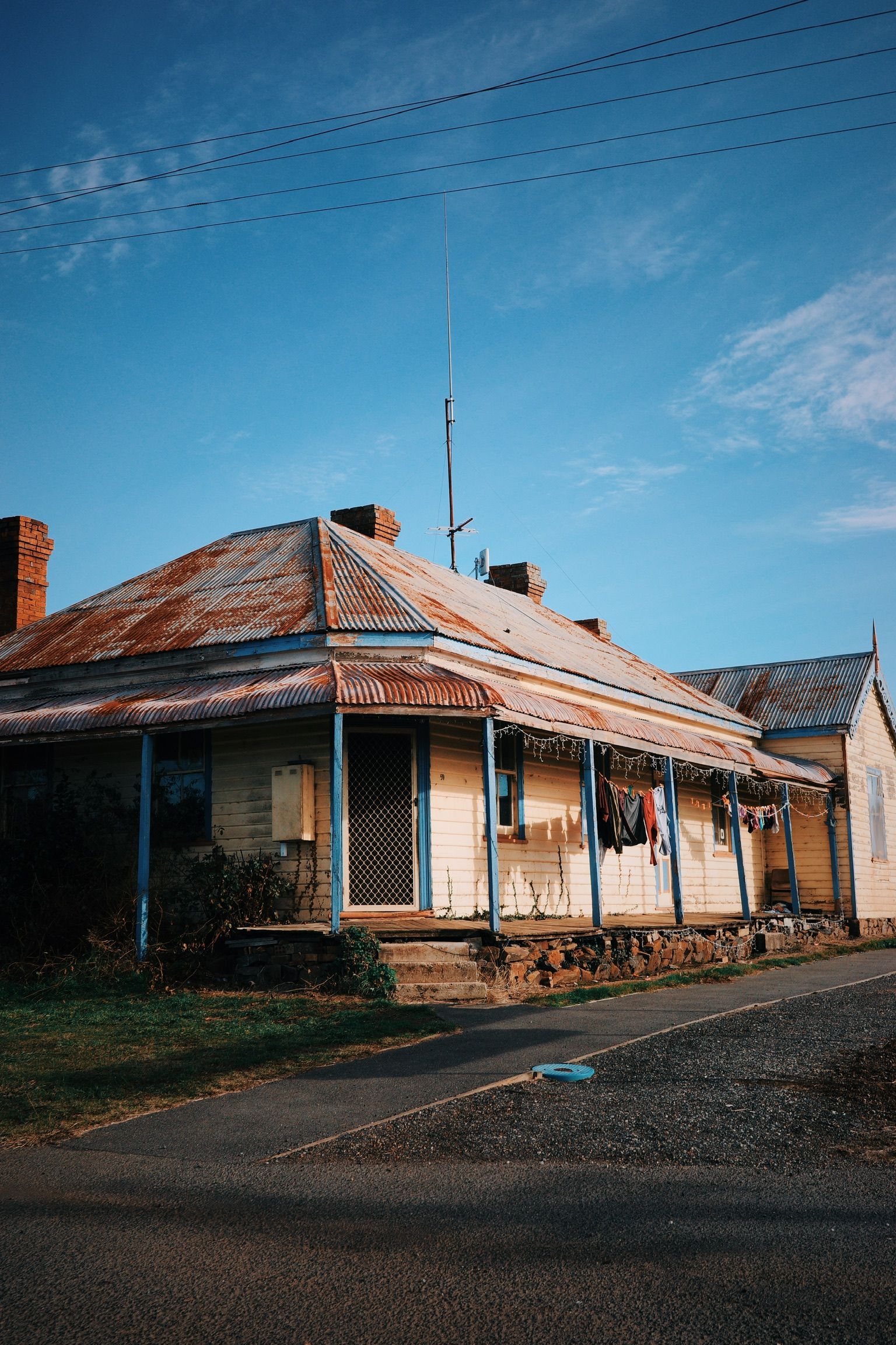 Historic weathered house with corrugated iron roof and blue trim beside rural road under blue sky