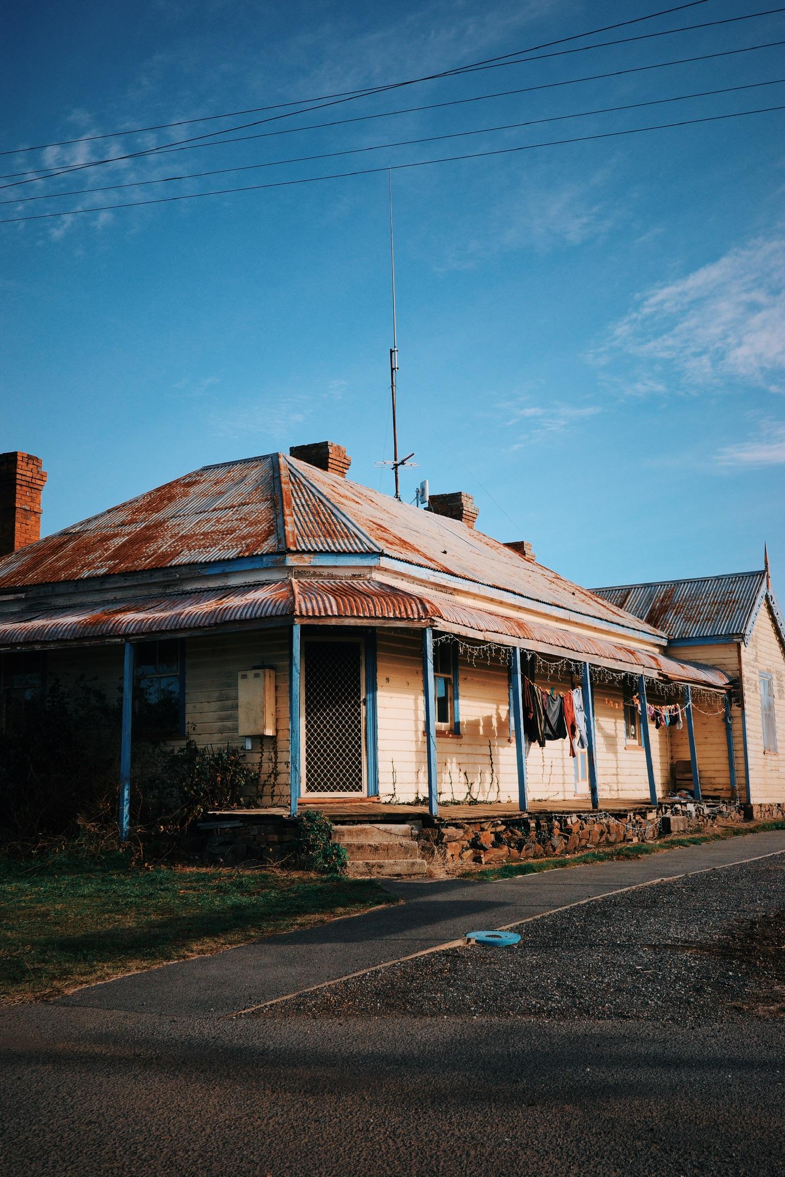 Historic weathered house with corrugated iron roof and blue trim beside rural road under blue sky