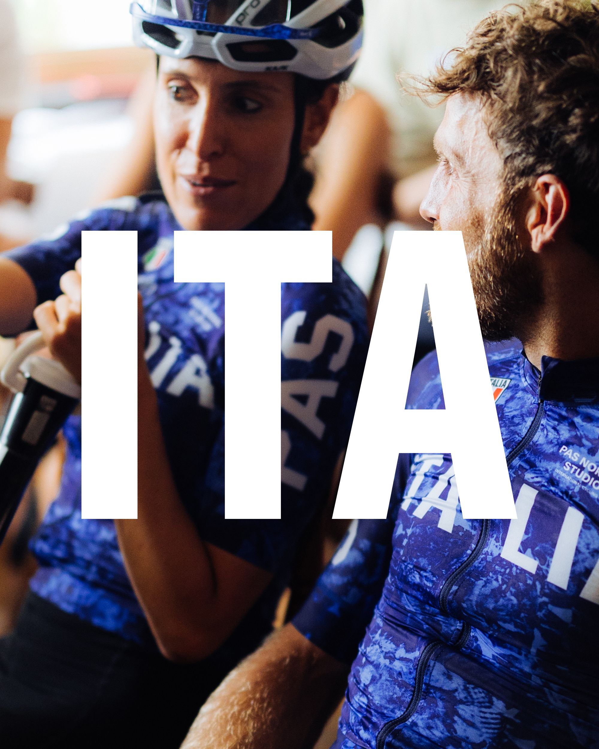 Two cyclists wearing blue Italia jerseys with white lettering, overlaid with large white "ITA" text
