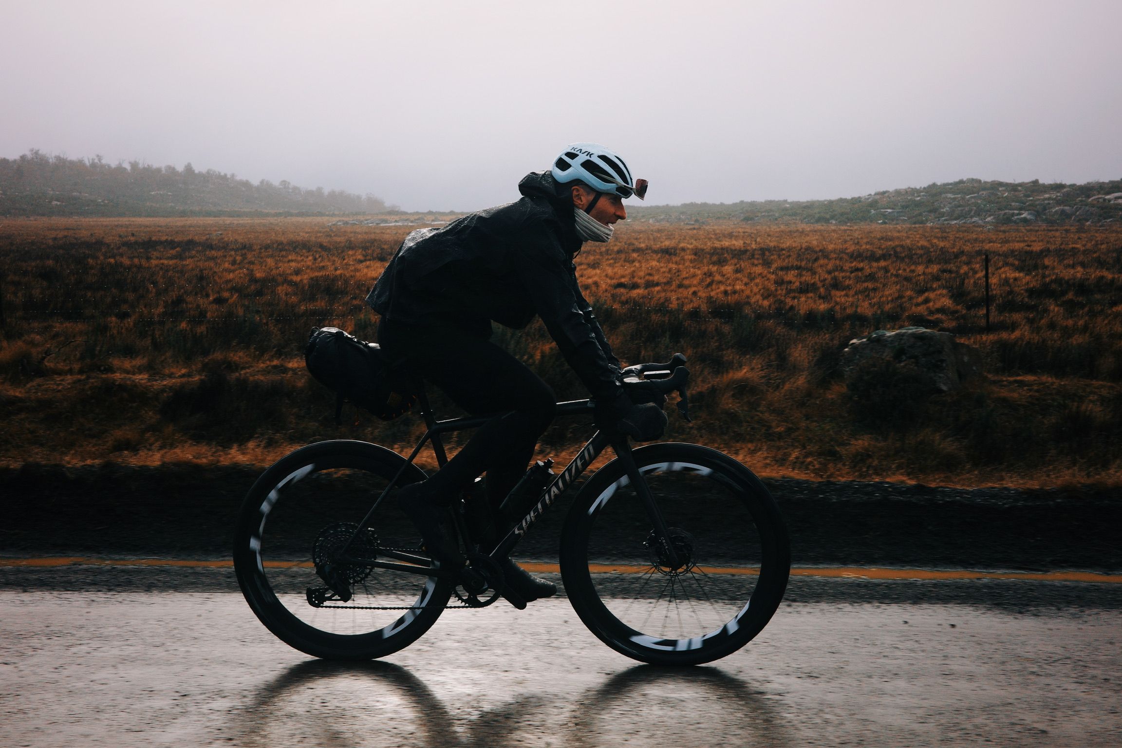Model wearing dark cycling apparel riding time trial bike on wet road through moorland landscape