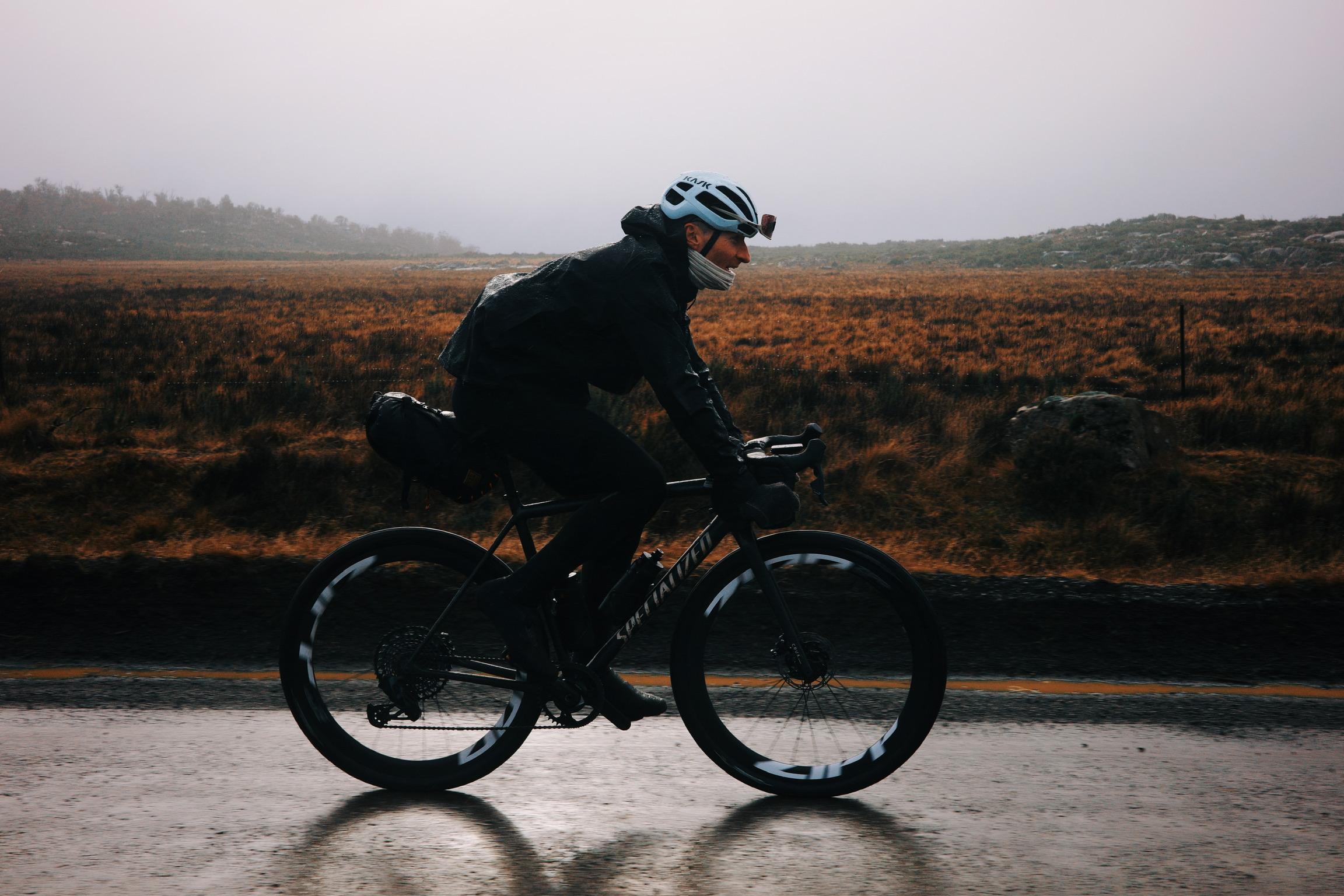 Model wearing dark cycling apparel riding time trial bike on wet road through moorland landscape
