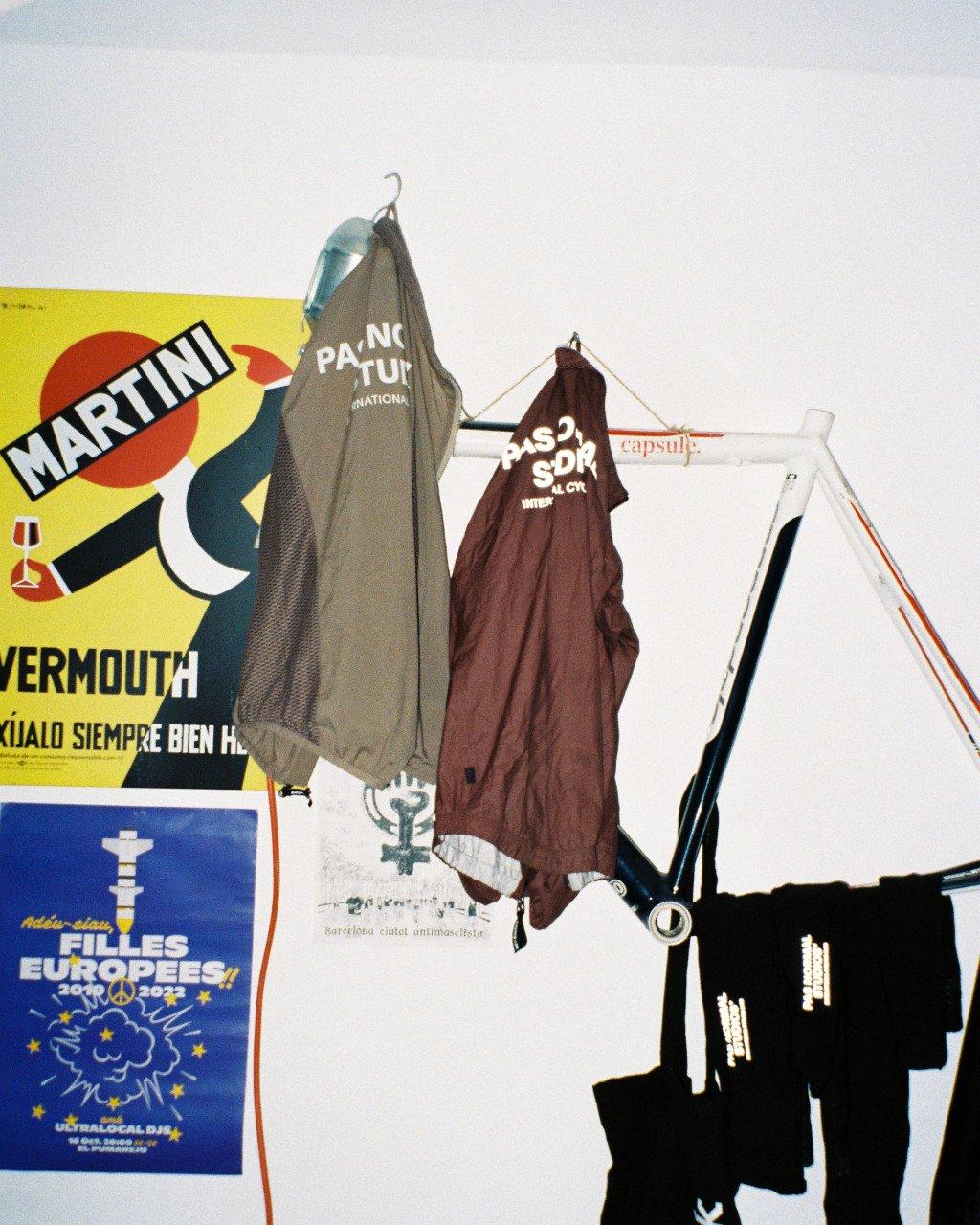Grey and burgundy cycling jerseys hanging in indoor space with vintage advertising posters on wall