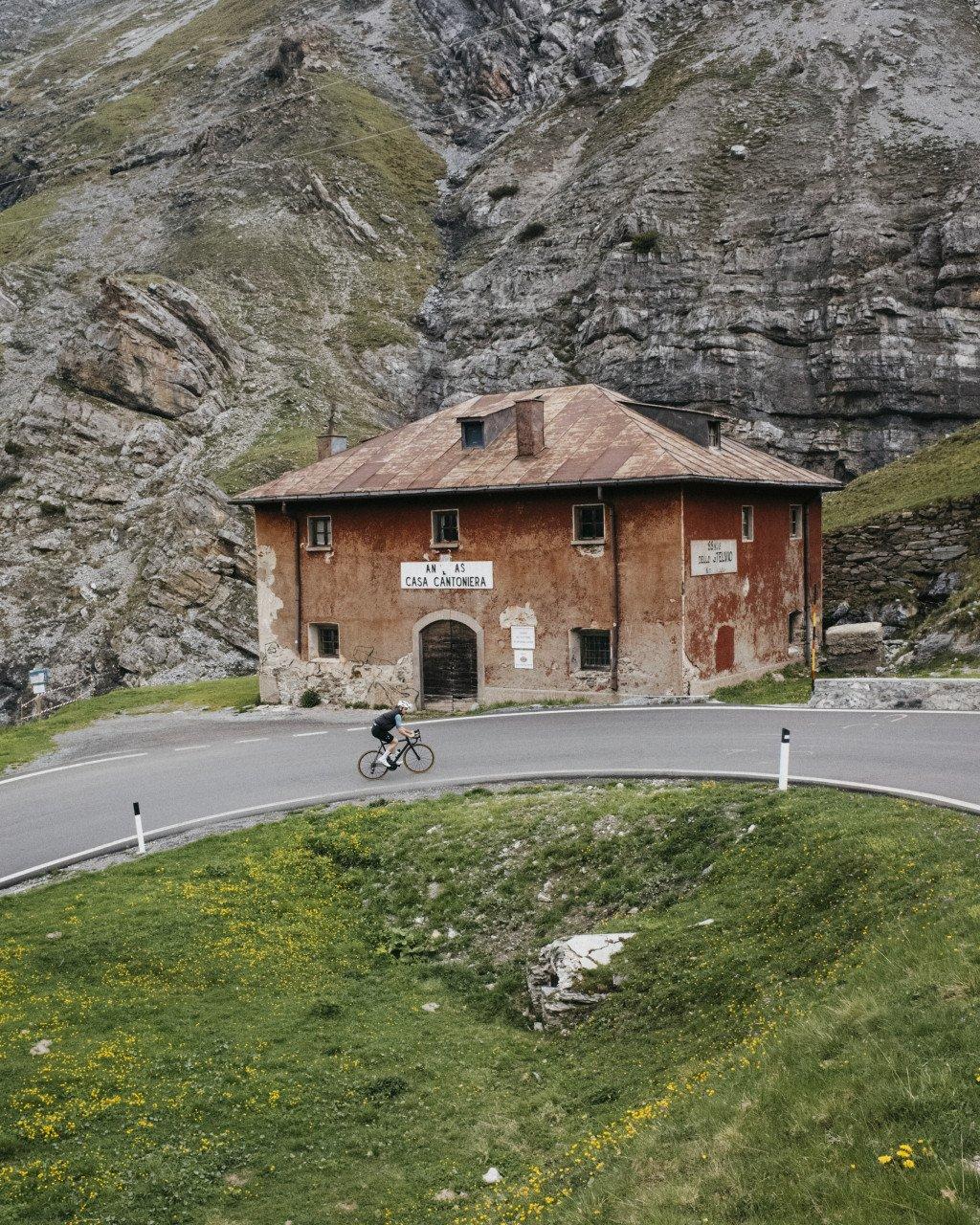 Cyclist on mountain road passing historic stone building with dramatic rocky cliffs backdrop