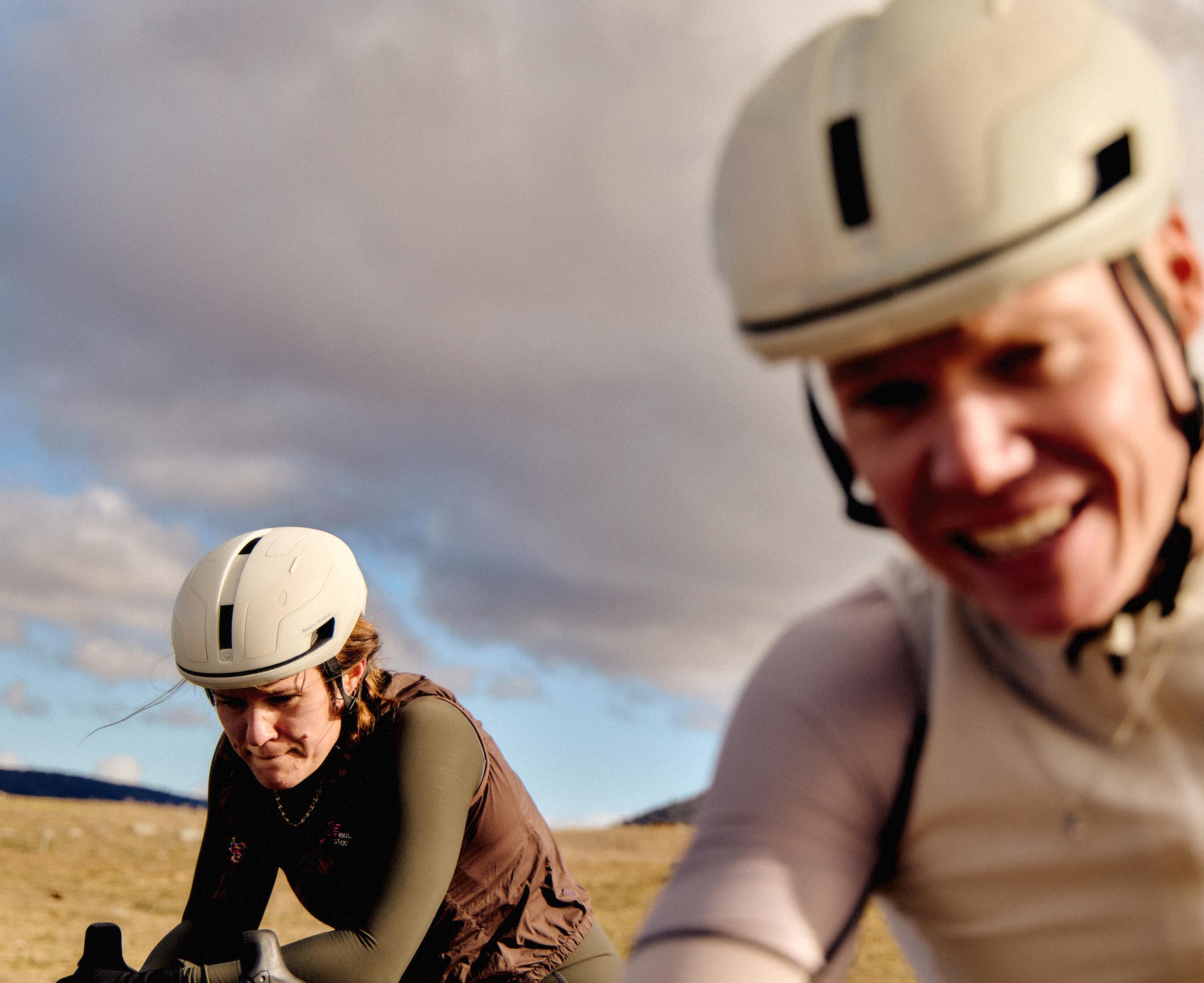 Two cyclists wearing white aero helmets and cycling apparel on outdoor terrain with cloudy sky
