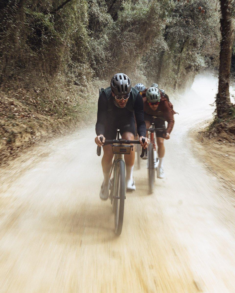 Models cycling on dusty gravel path through wooded terrain wearing dark cycling apparel and helmets