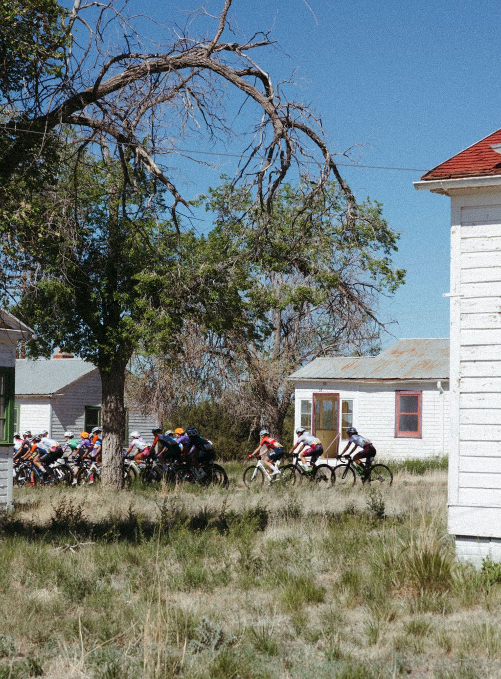 Cyclists racing past rural white buildings with red trim during Tour of the Gila