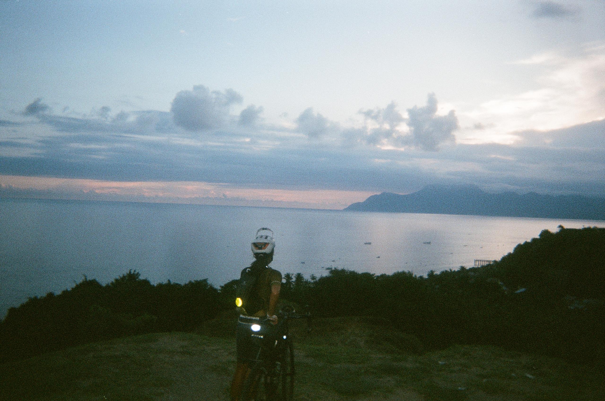 Model on bike at dusk overlooking coastal waters with mountains and cloudy sky