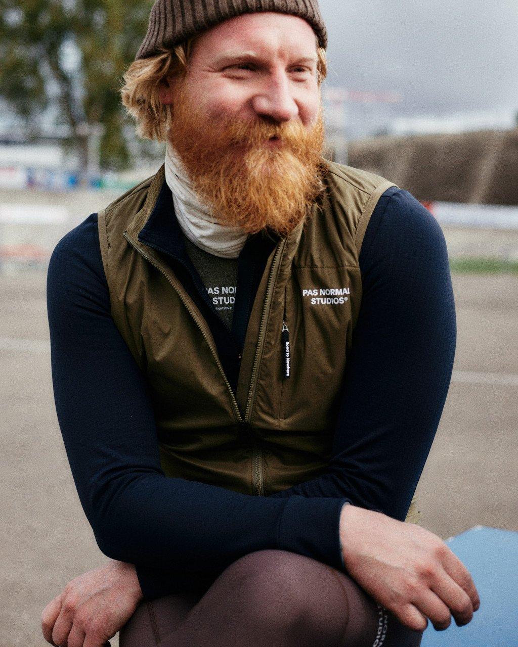 Model wearing olive green vest over navy long sleeve base layer with brand logo, outdoors in urban setting