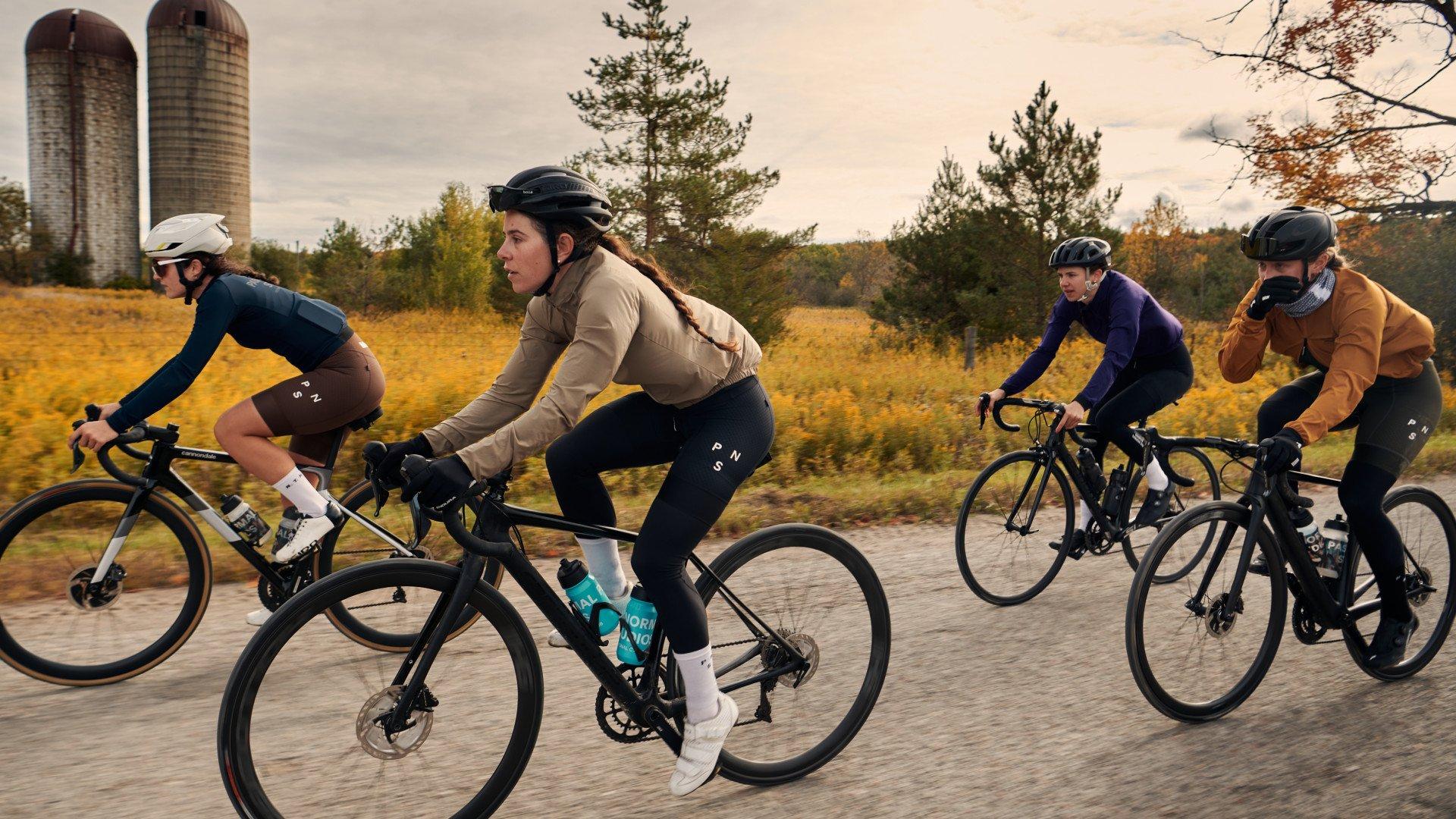 Four cyclists riding road bikes on gravel path through autumn countryside with farm silos in background