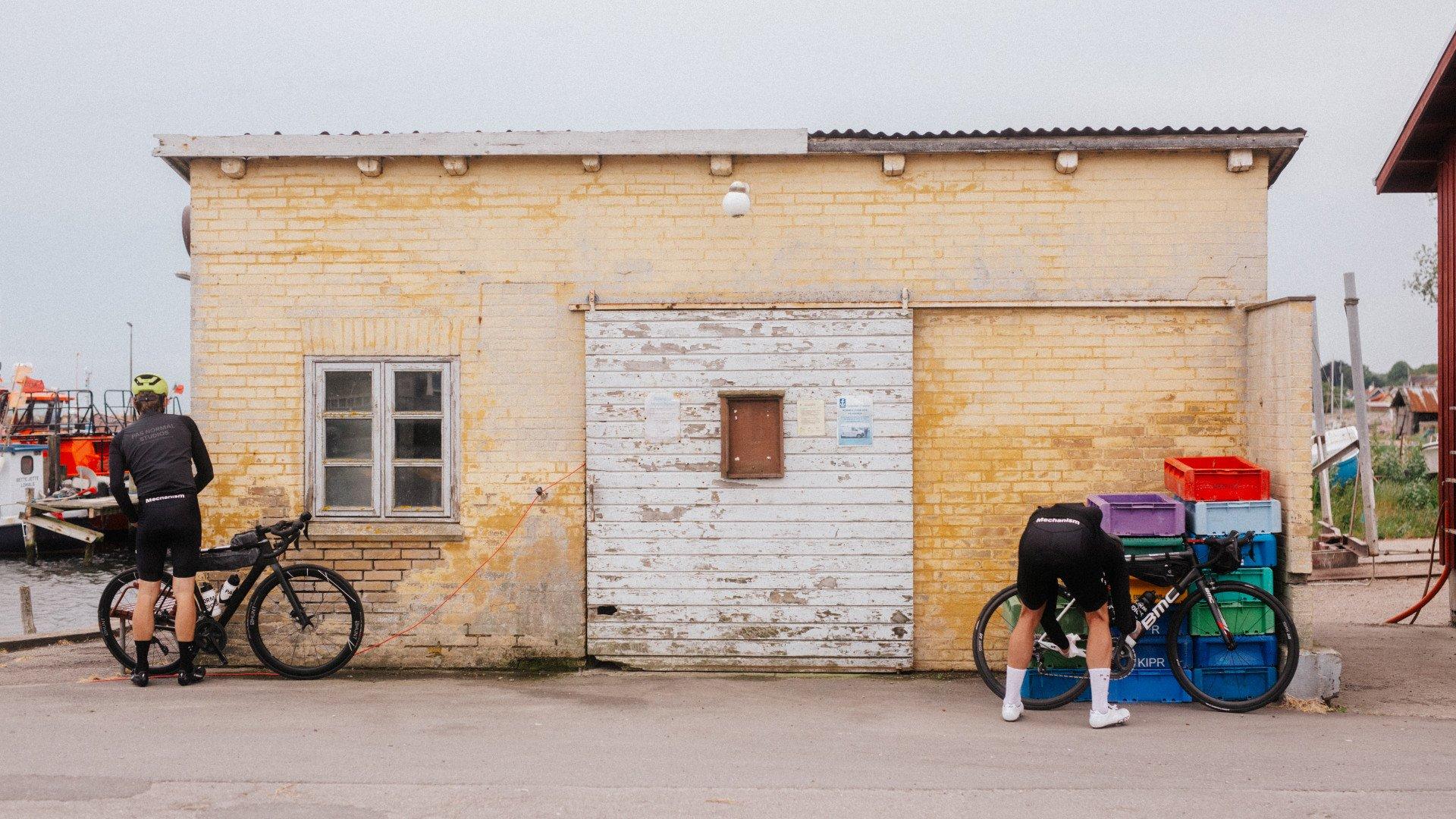 Two cyclists in black cycling gear with bikes beside weathered coastal building with colorful crates