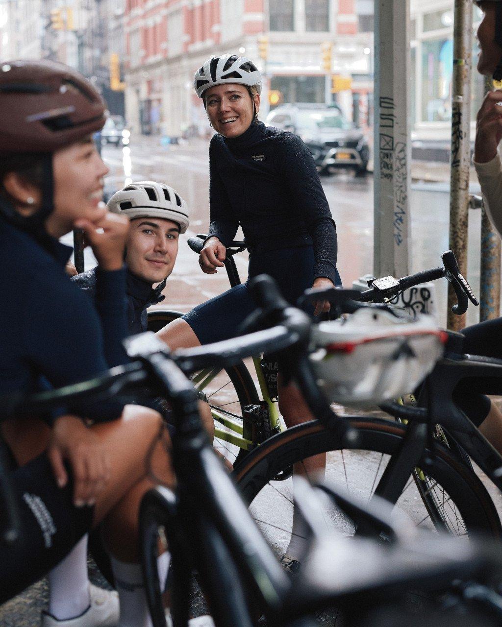 Group of cyclists with bikes on city street, woman in black long sleeve jersey smiling in background