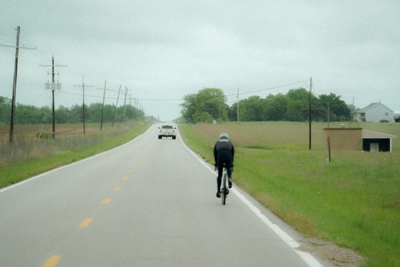 Cyclist in dark clothing riding on rural road with approaching vehicle