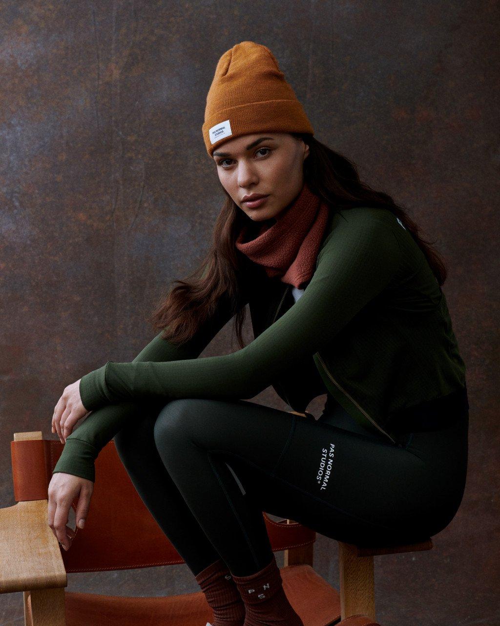 Model wearing green long-sleeve top, black tights with leg text, orange beanie, and burgundy scarf in studio