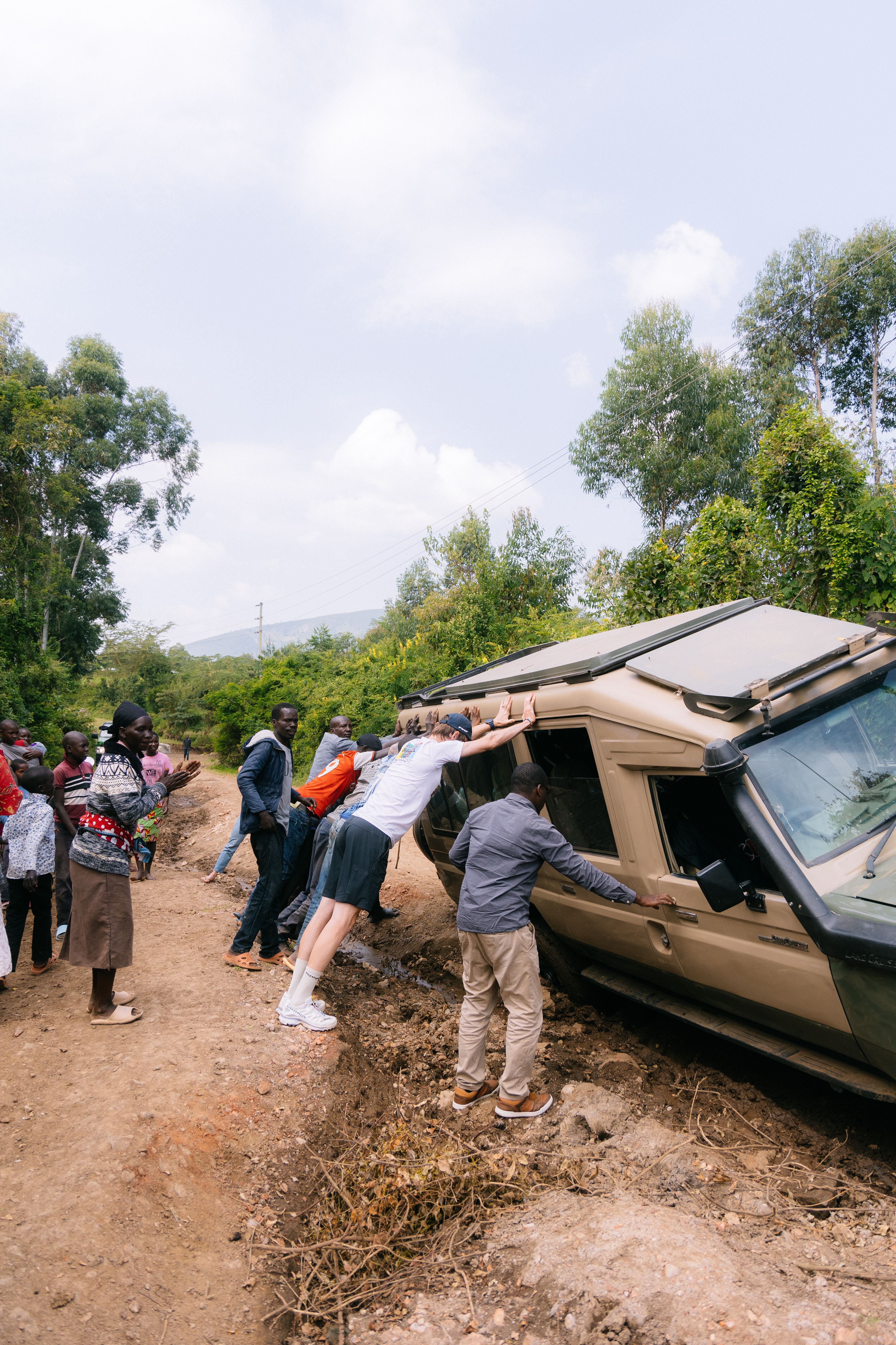 Group of people pushing stuck vehicle on rural dirt road in mountainous landscape