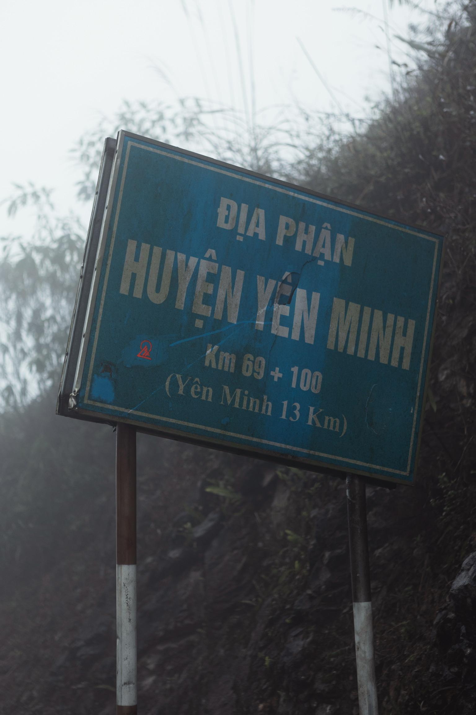 Blue road sign in Vietnamese showing distance to Yen Minh district against misty mountain backdrop