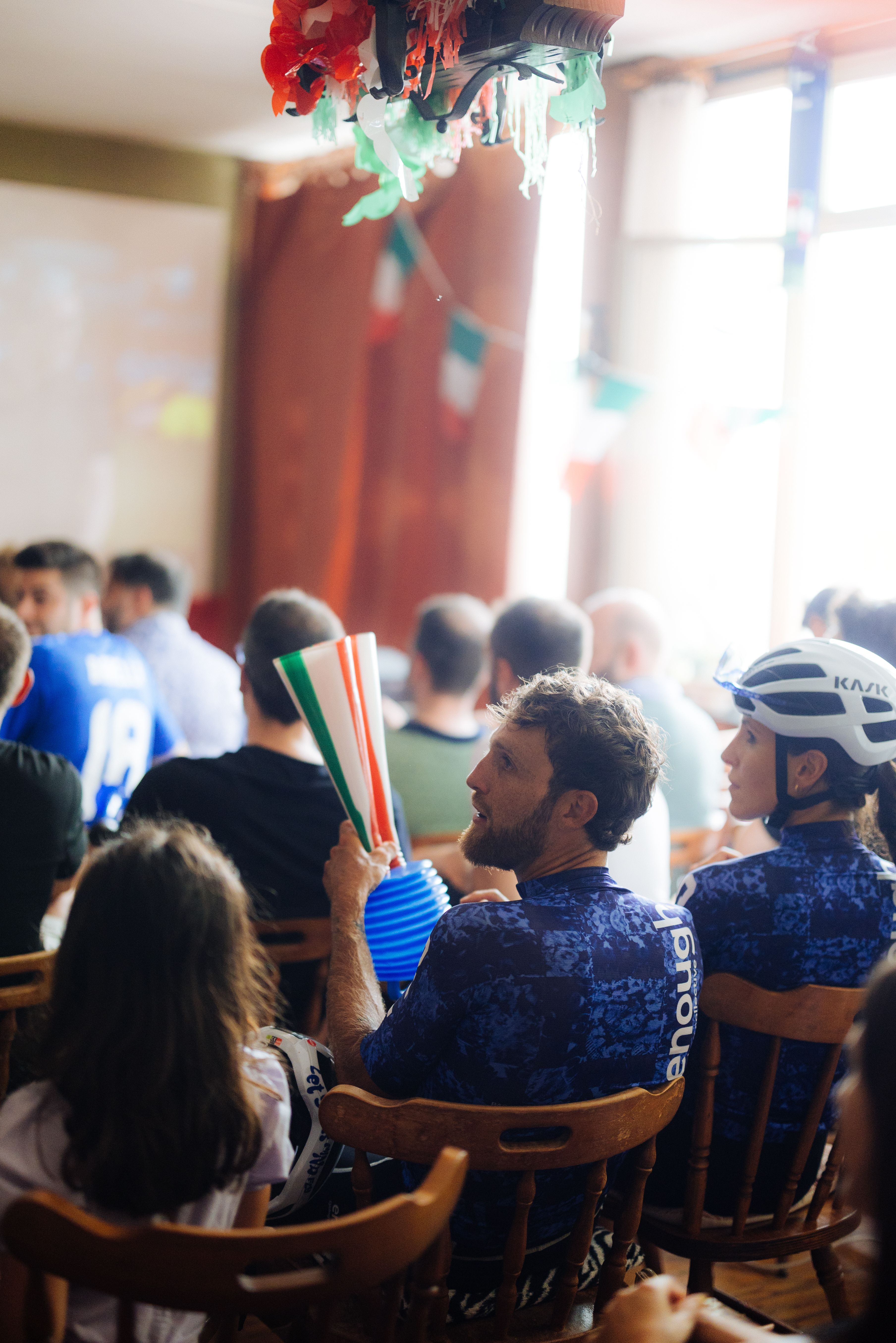 Cyclists in blue patterned jerseys watching event indoors, one holding Italian flag