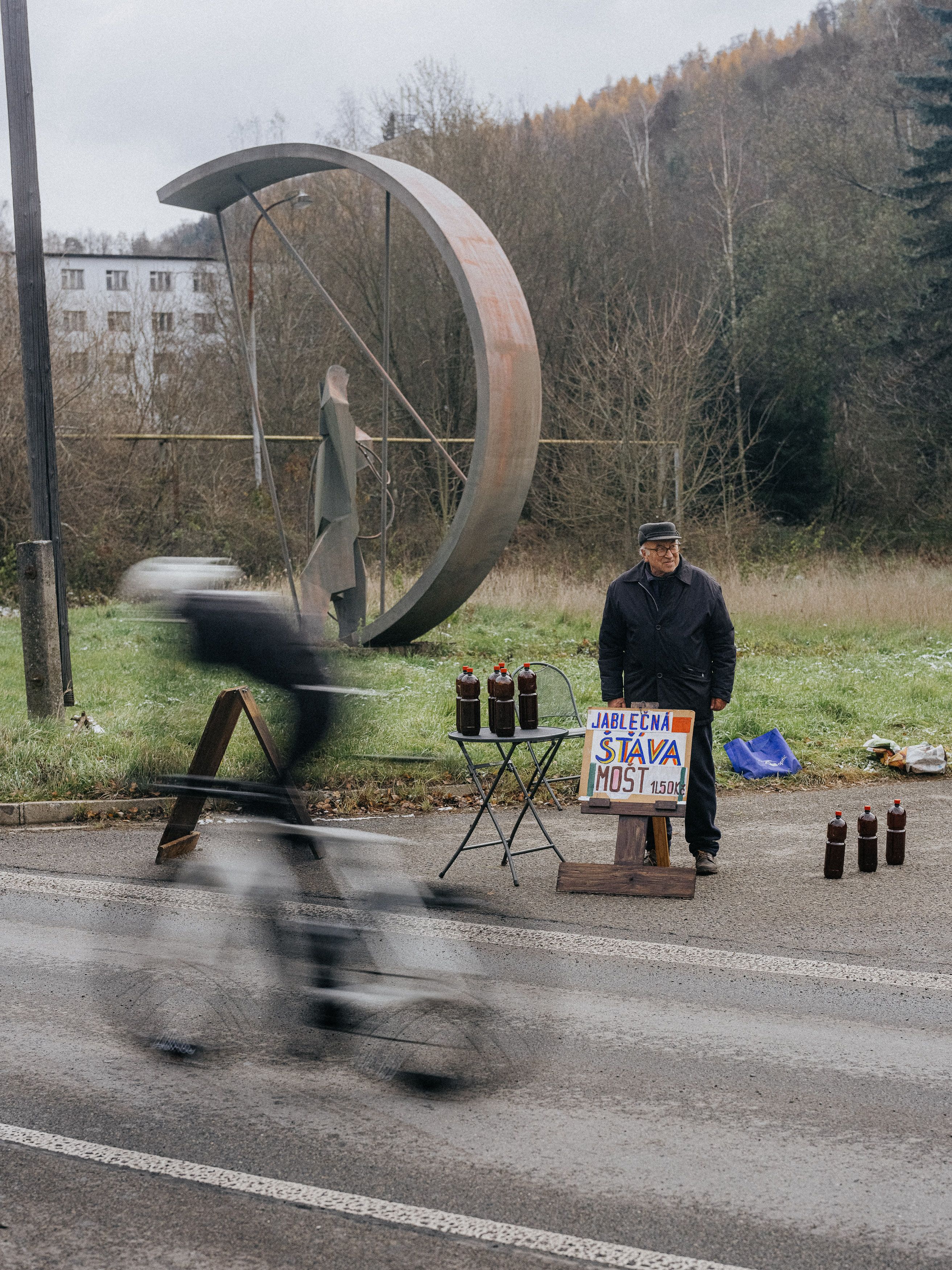 Cyclist in motion blur passing roadside vendor near modern sculpture in rural landscape
